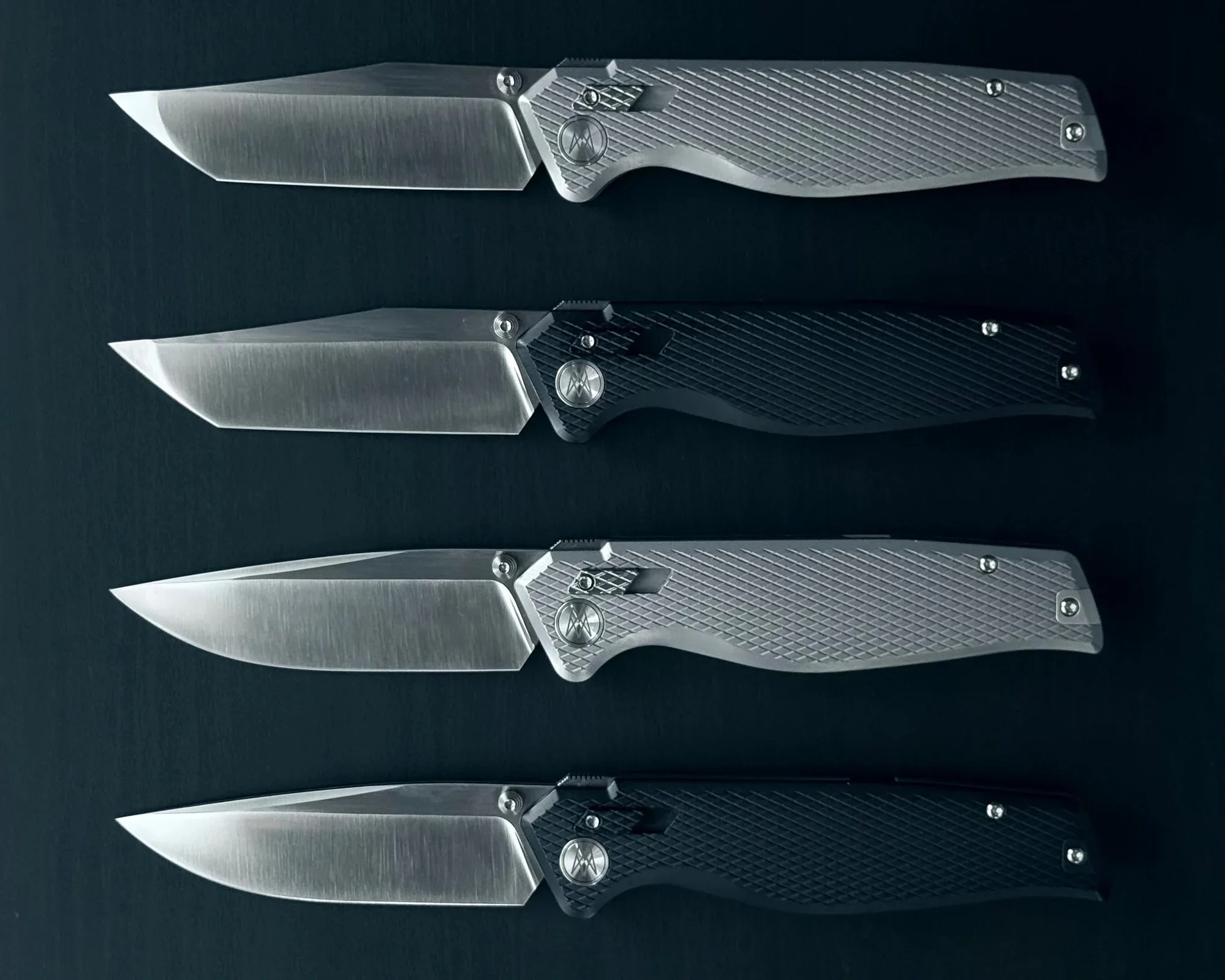 Four folding knives with textured handles, two silver and two black, arranged horizontally on a dark surface.