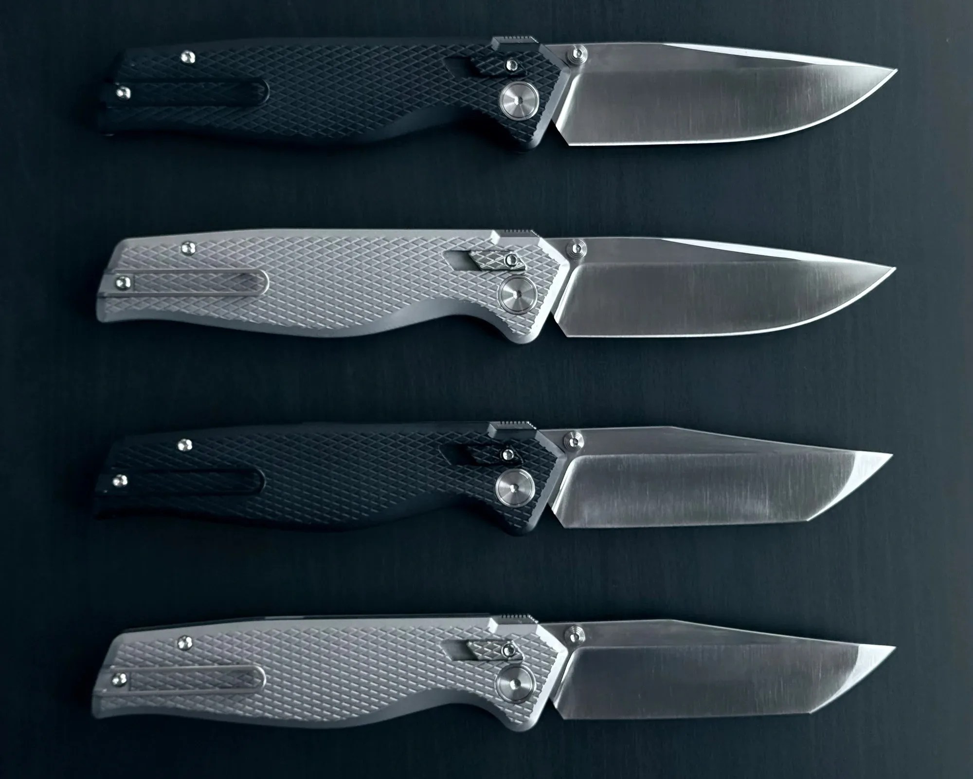 Four folding knives with textured handles, two black and two silver, arranged in alternating colors on a dark surface.