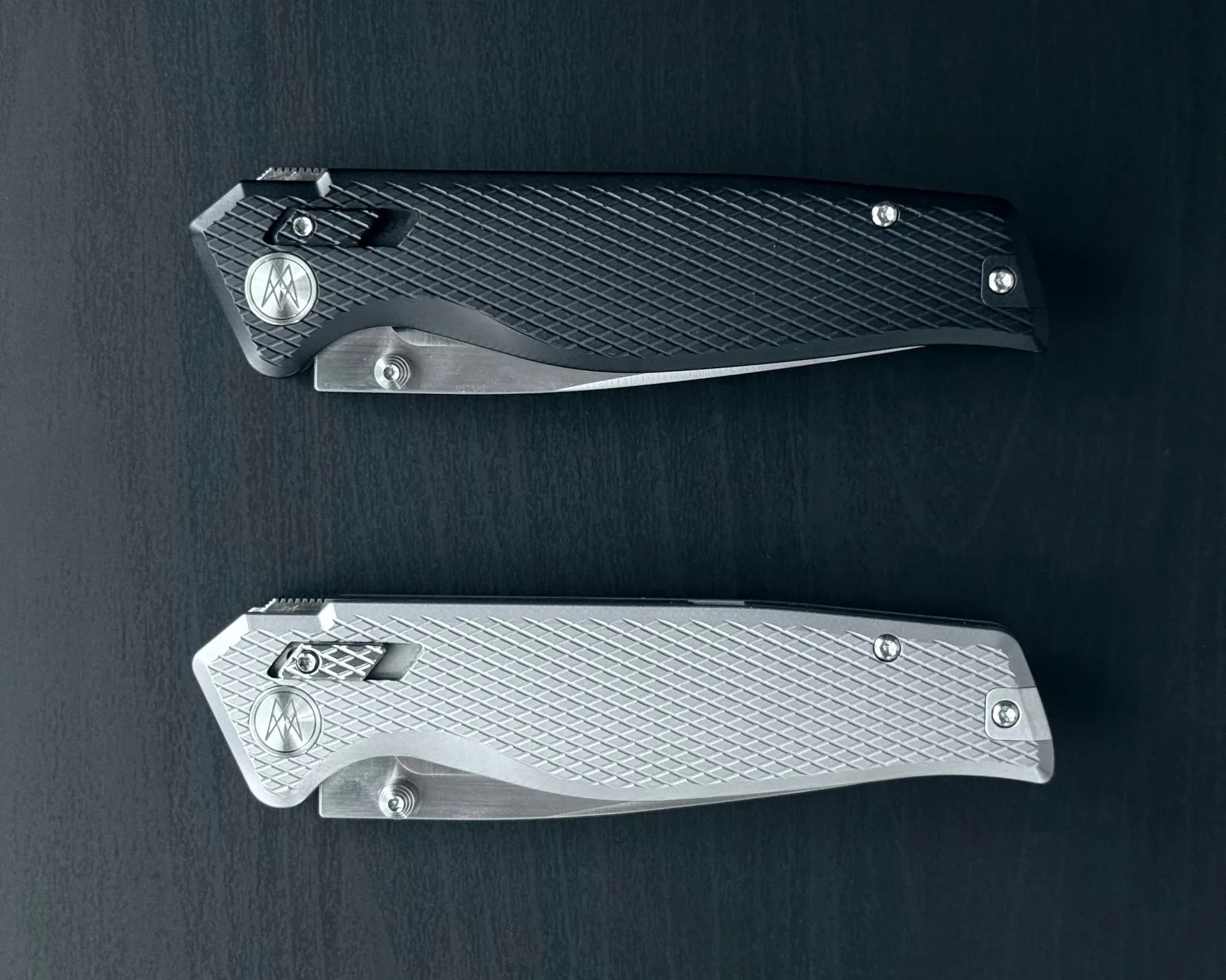 Two closed folding knives with textured handles, one black and one silver, placed on a dark surface.