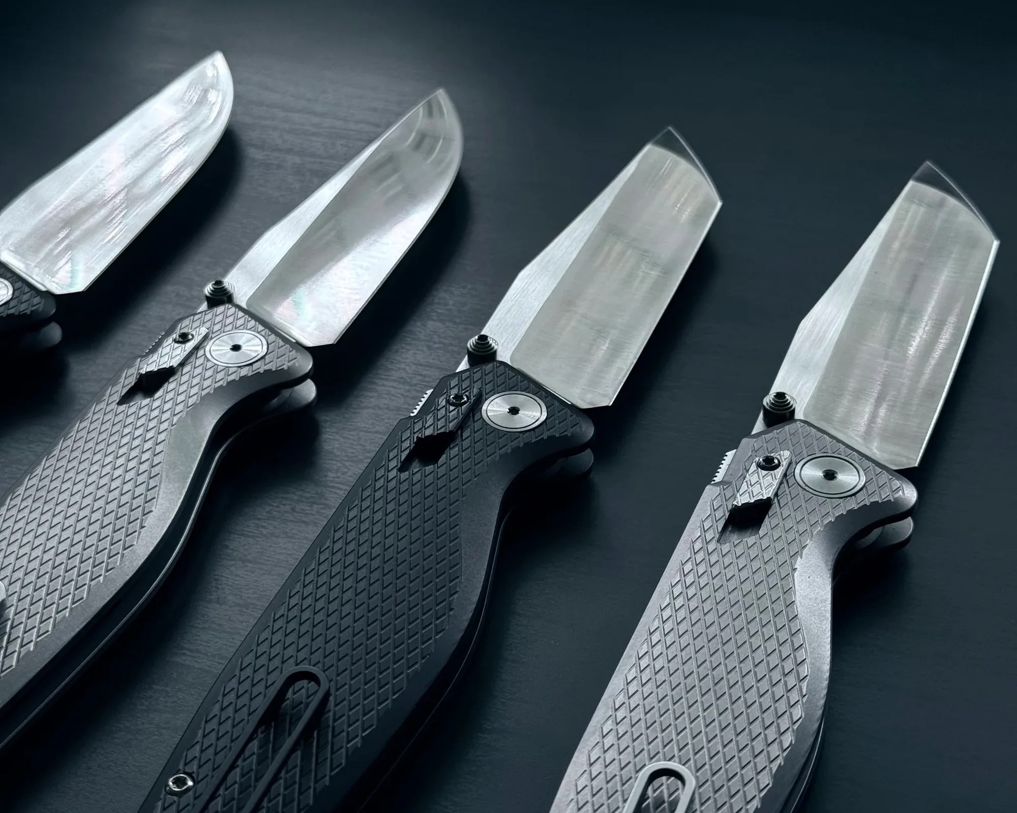 Four folding knives with textured black handles and wide, polished stainless steel blades arranged on a dark surface.