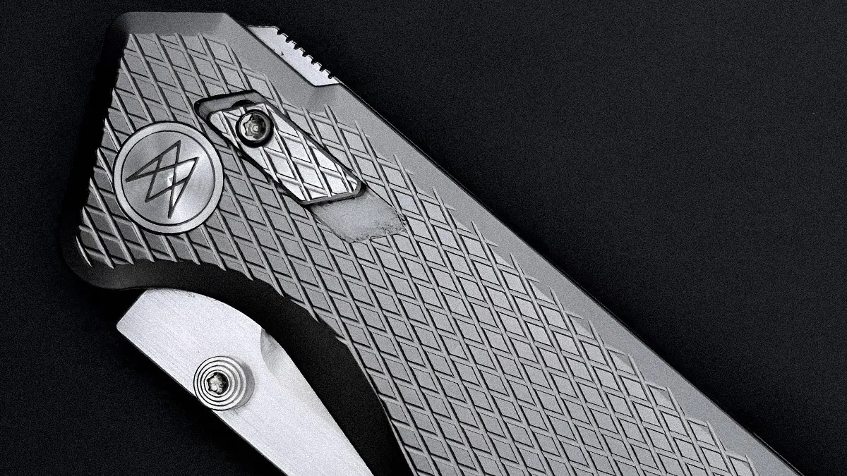 Close-up of a silver folding knife handle with diamond-pattern texture and a circular logo on a black background.