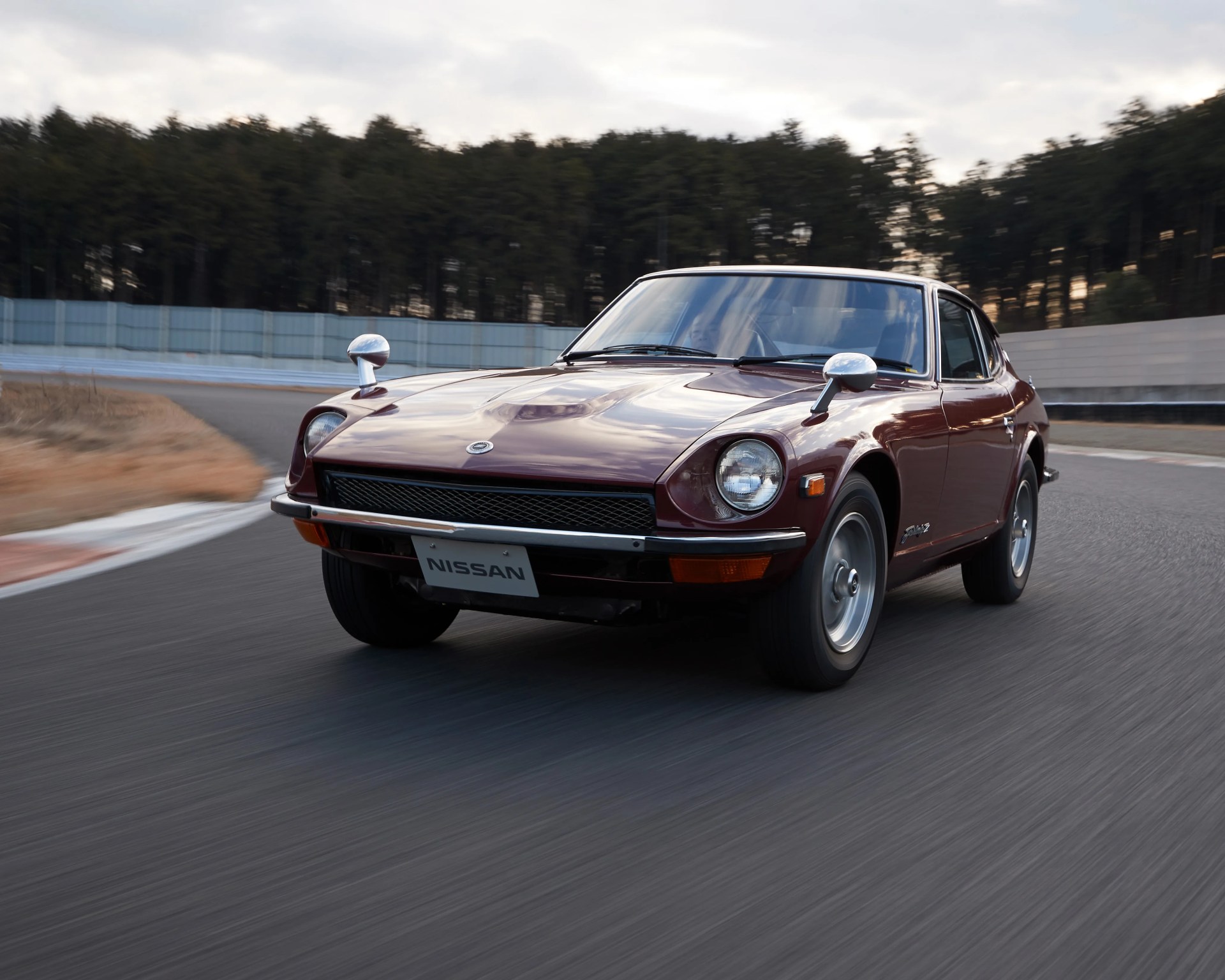 Maroon vintage Nissan sports car driving on a racetrack with trees in the background.