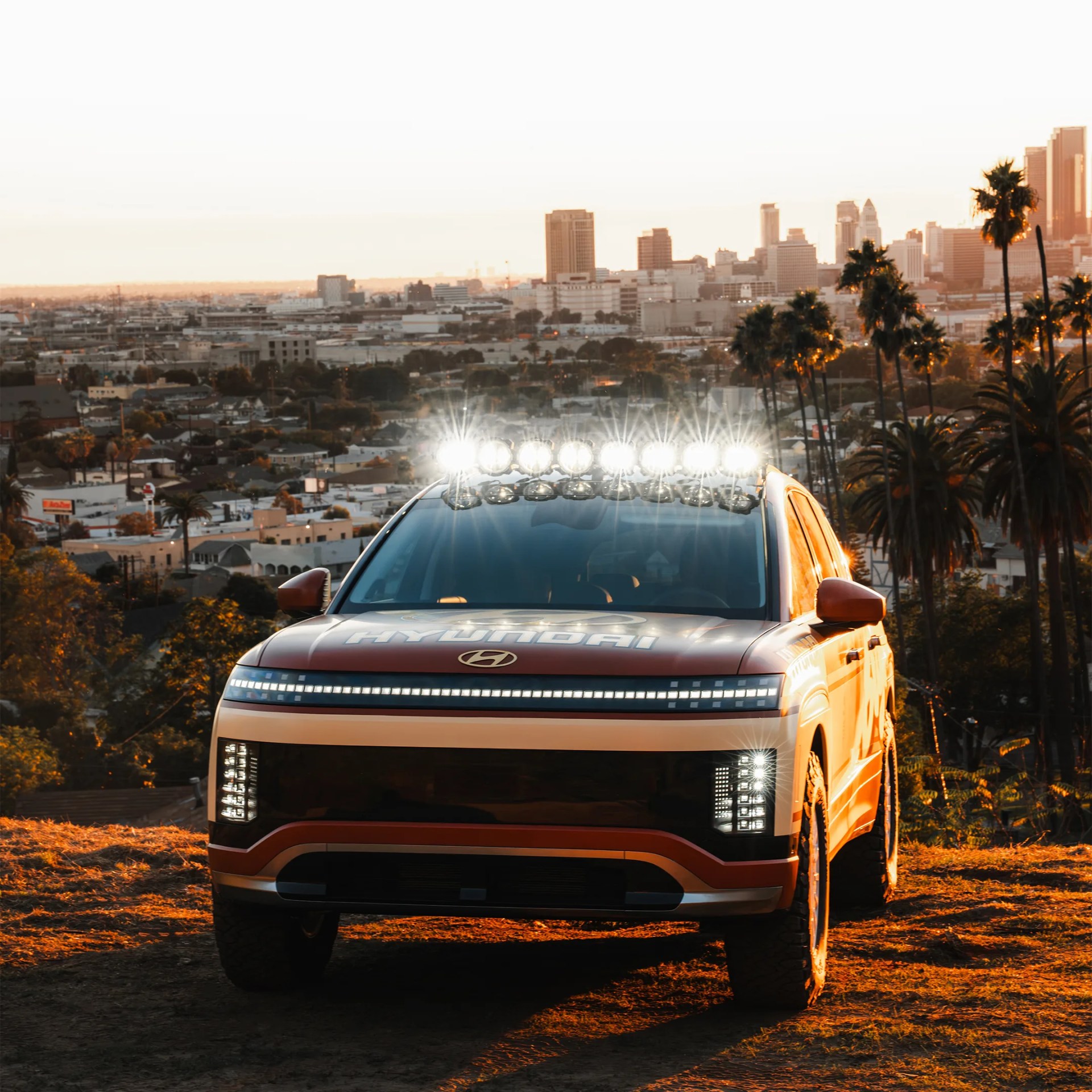 Red Hyundai SUV with roof-mounted off-road lights illuminated, parked on a hill overlooking a cityscape at sunset.