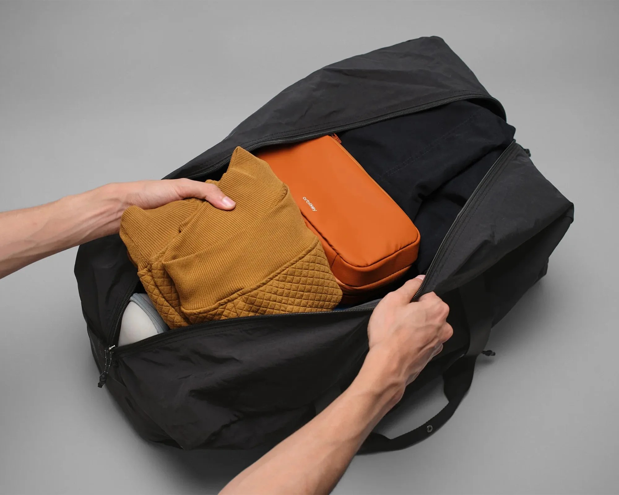 Black duffel bag being packed with a folded brown quilted sweater and an orange packing cube.