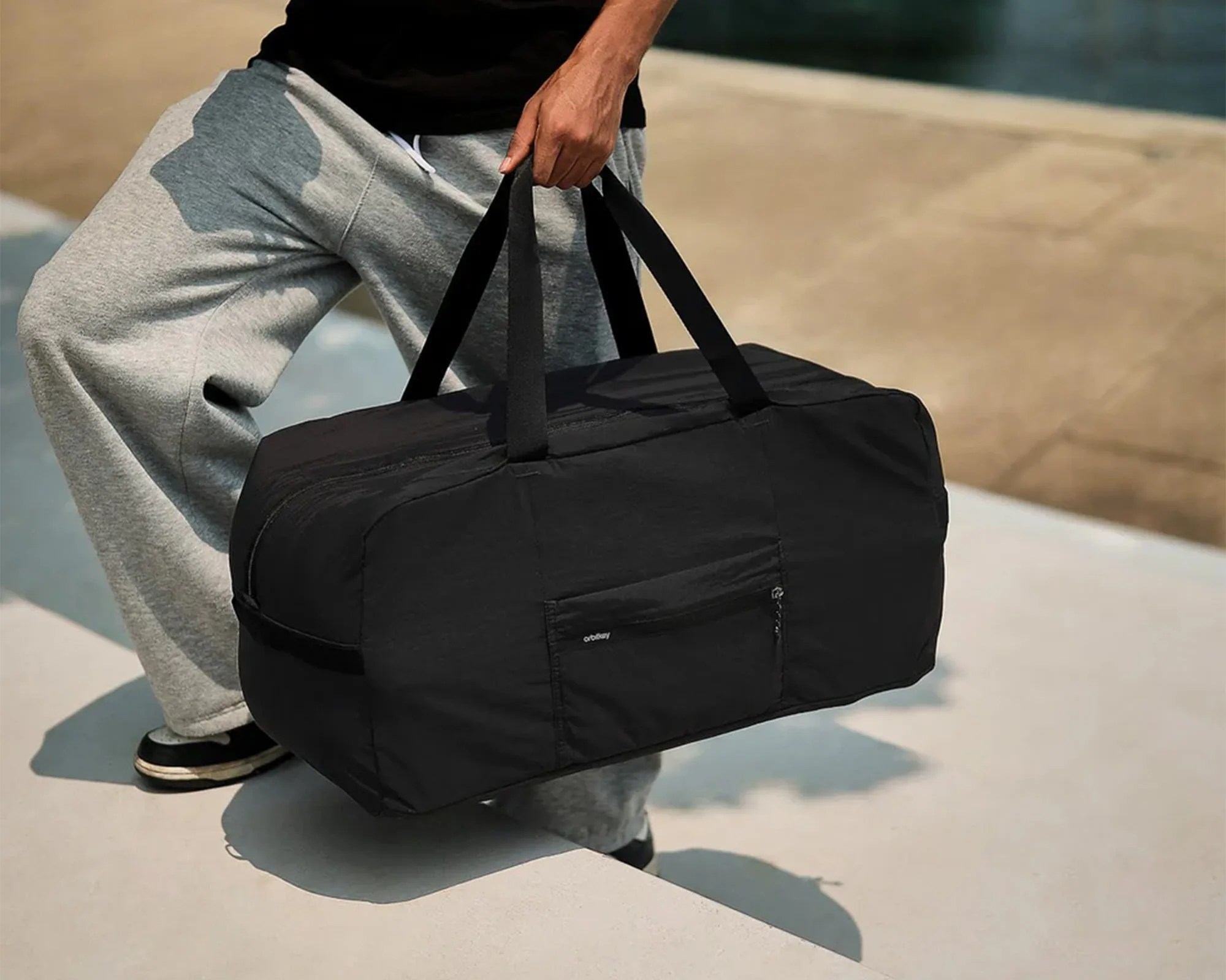 Black rectangular duffel bag with dual handles held by a person wearing gray sweatpants and black shoes.
