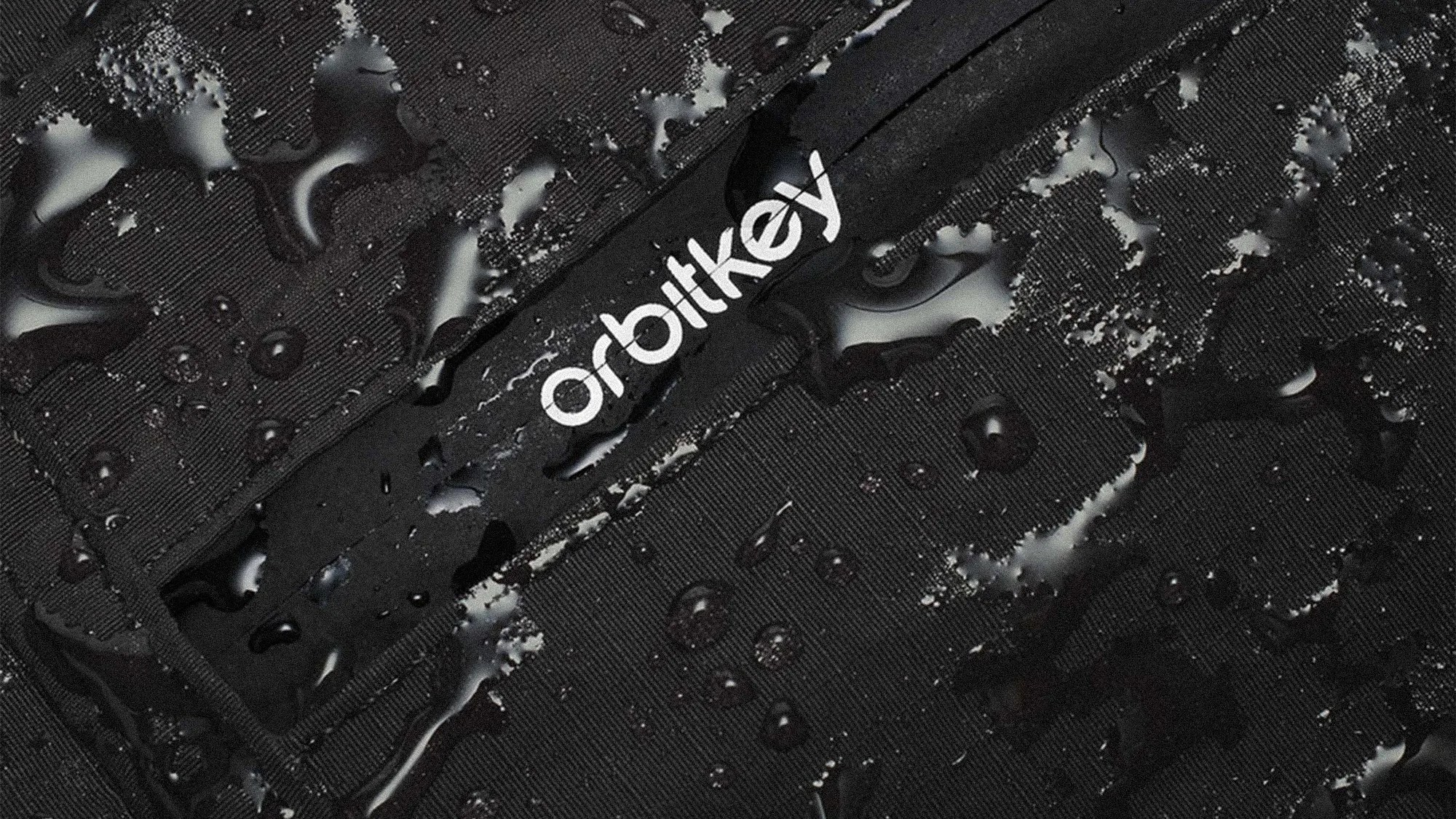 Close-up of black fabric with water droplets and a white "orbitkey" logo on a waterproof zipper.
