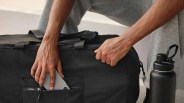 Person placing a silver smartphone into the front zippered pocket of a black duffel bag with a black water bottle nearby.