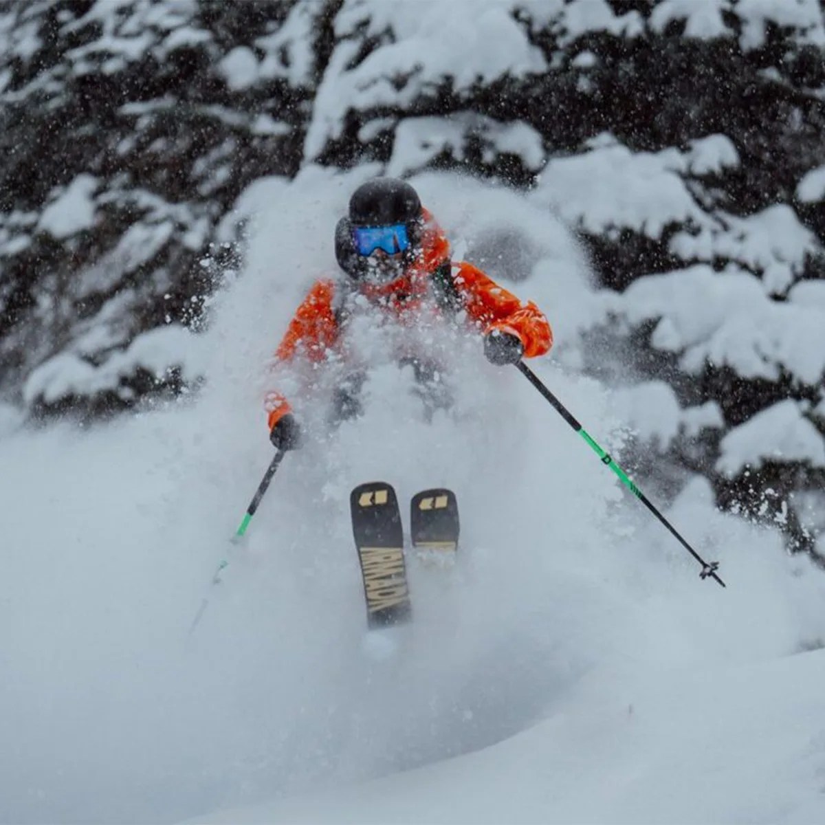 Skier in an orange jacket and blue goggles skiing through deep powder snow with snow-covered trees in the background.