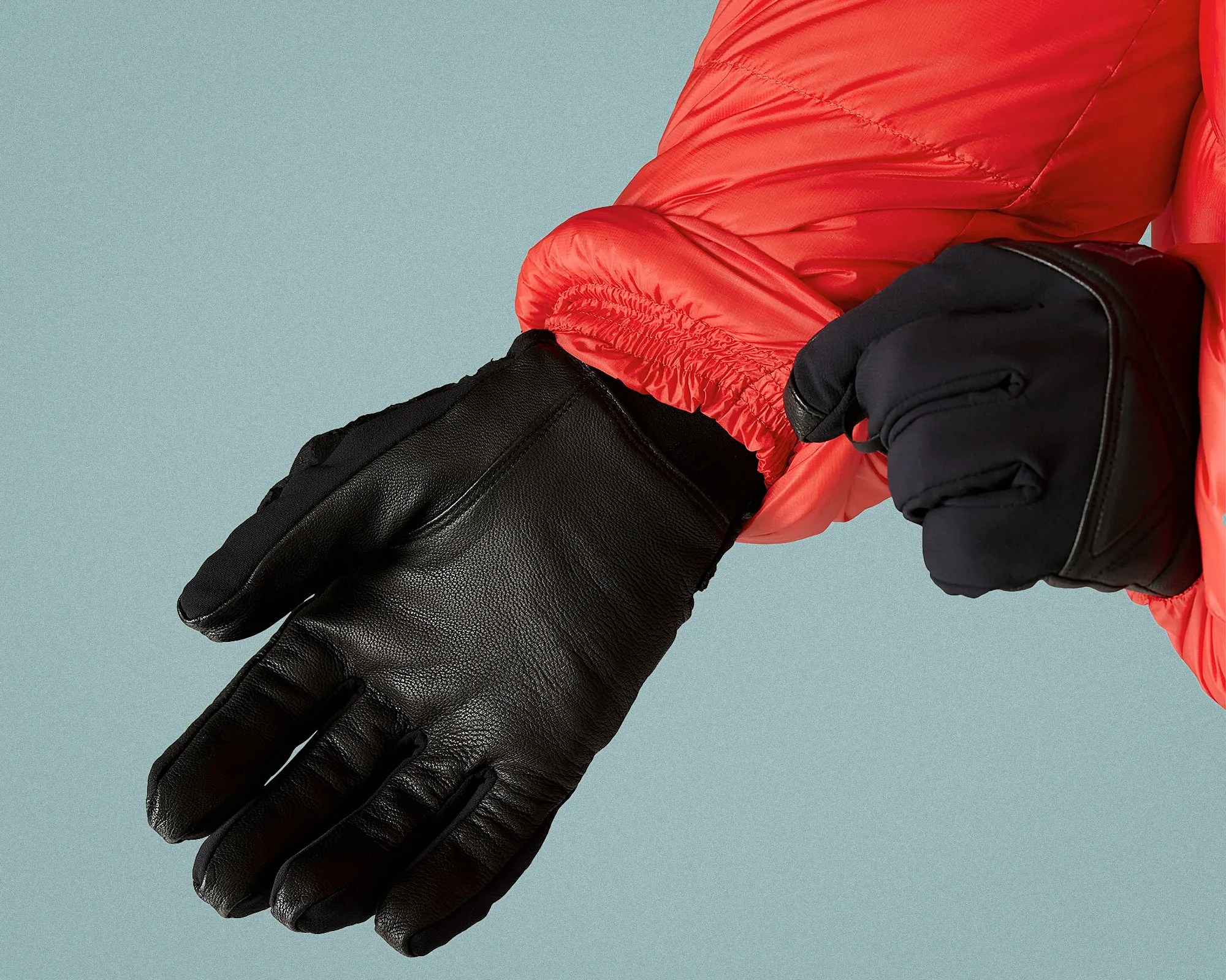 Pair of black leather and fabric gloves worn with a bright red jacket against a light blue background.