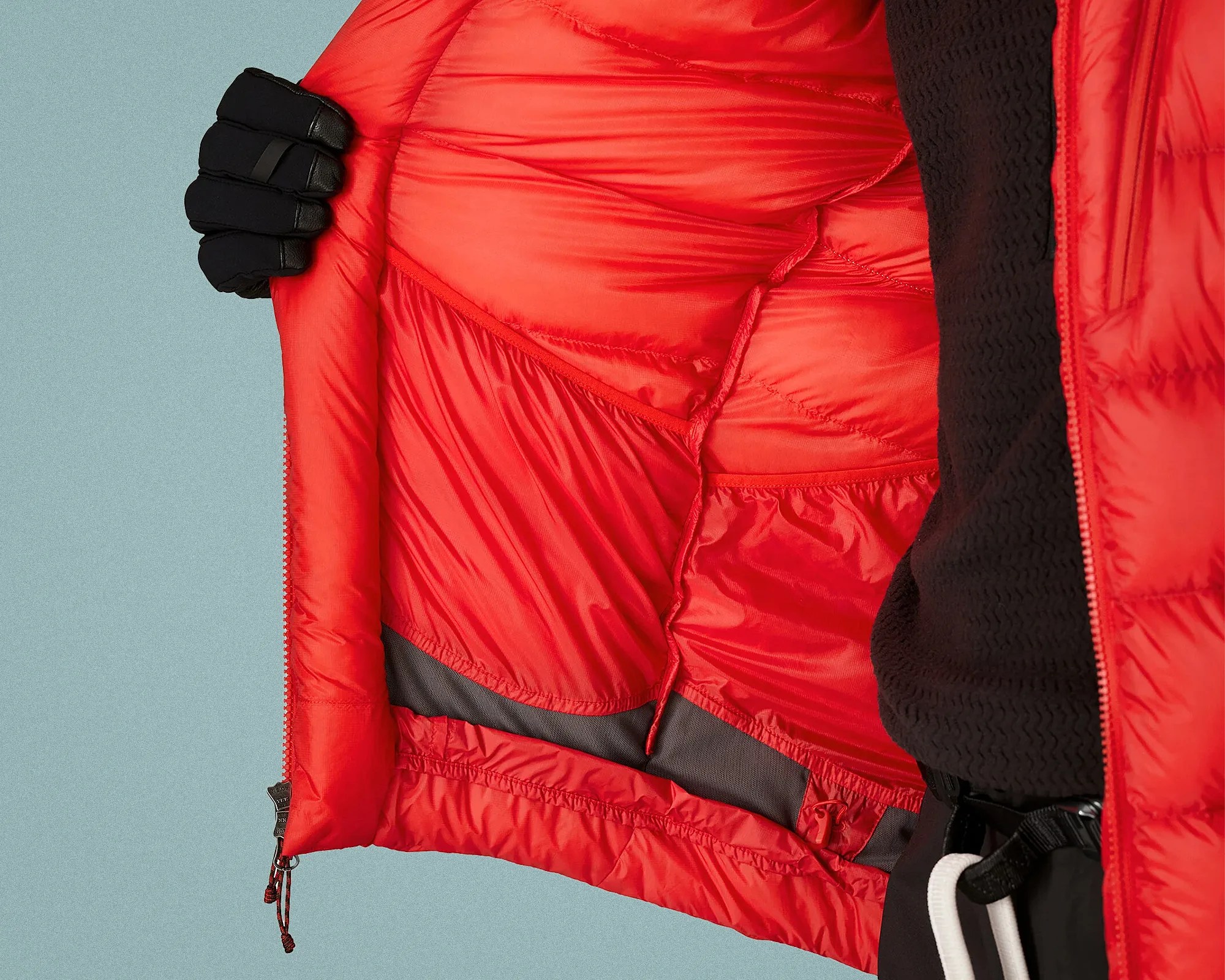 Inside view of a red insulated jacket showing internal stitching and a black mesh pocket, held open by a hand in a black glove.