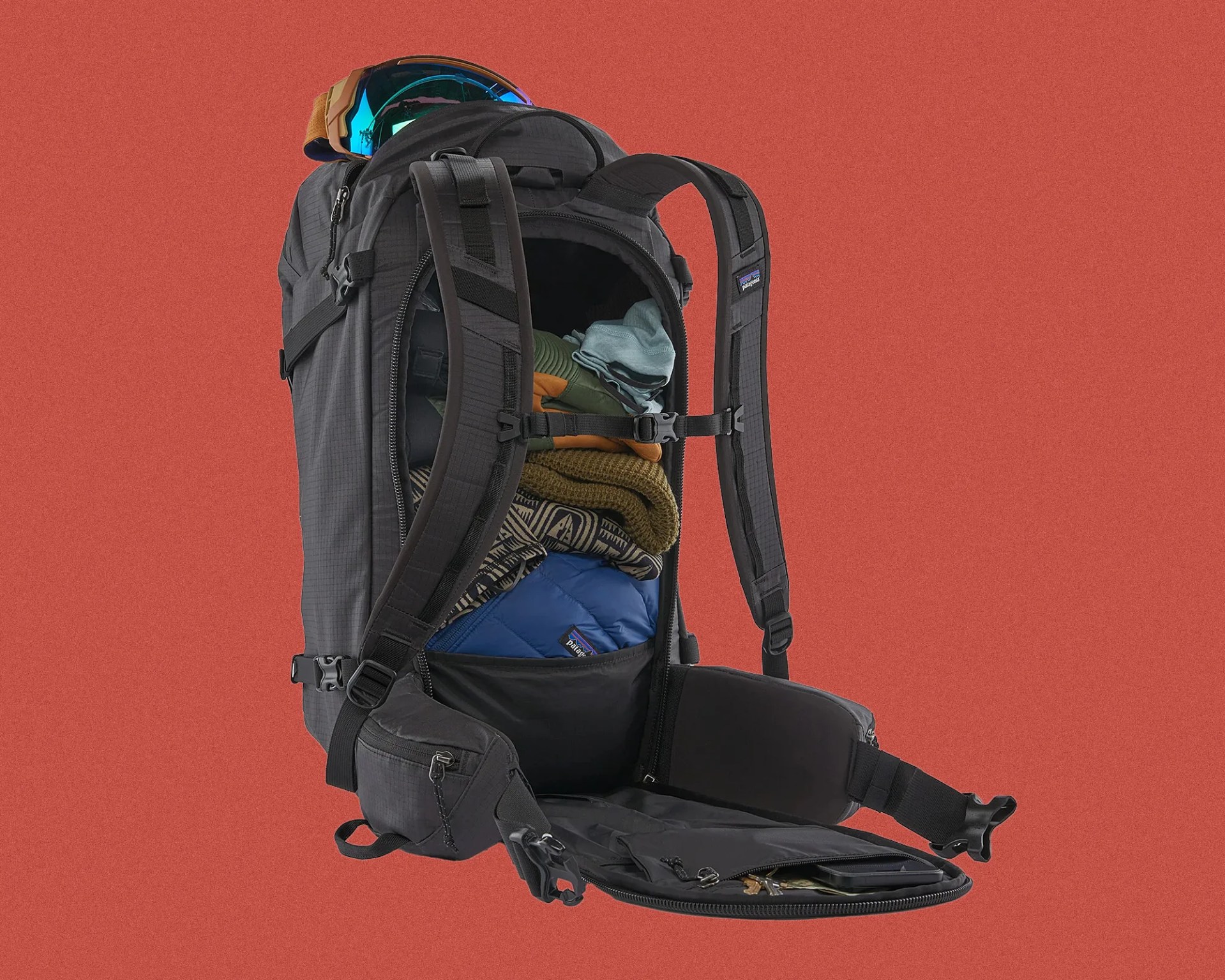 Black Patagonia backpack with padded straps and hip belt, open to show packed clothes and ski goggles on top.