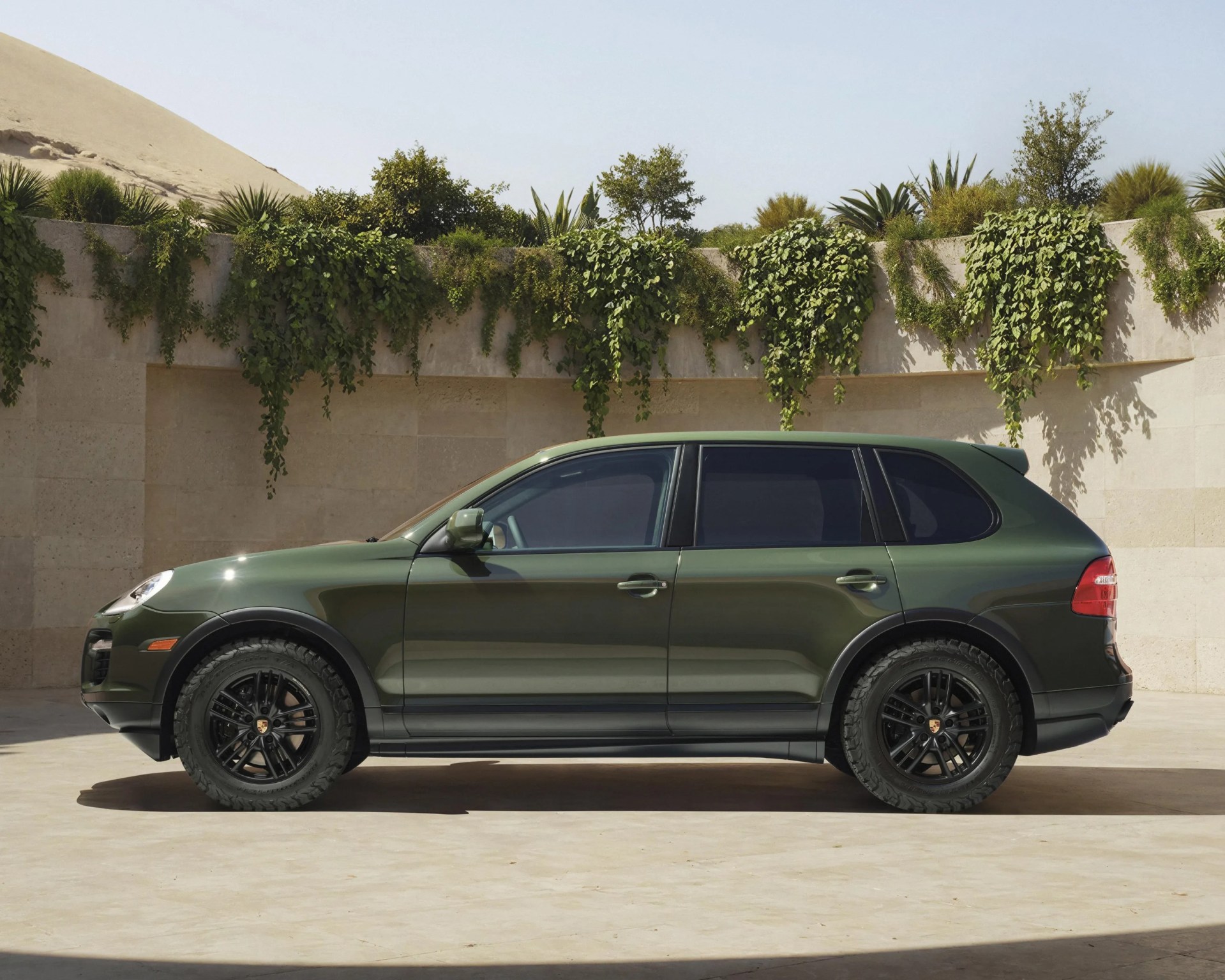 Dark green Porsche SUV with black wheels parked in front of a beige stone wall with hanging greenery.