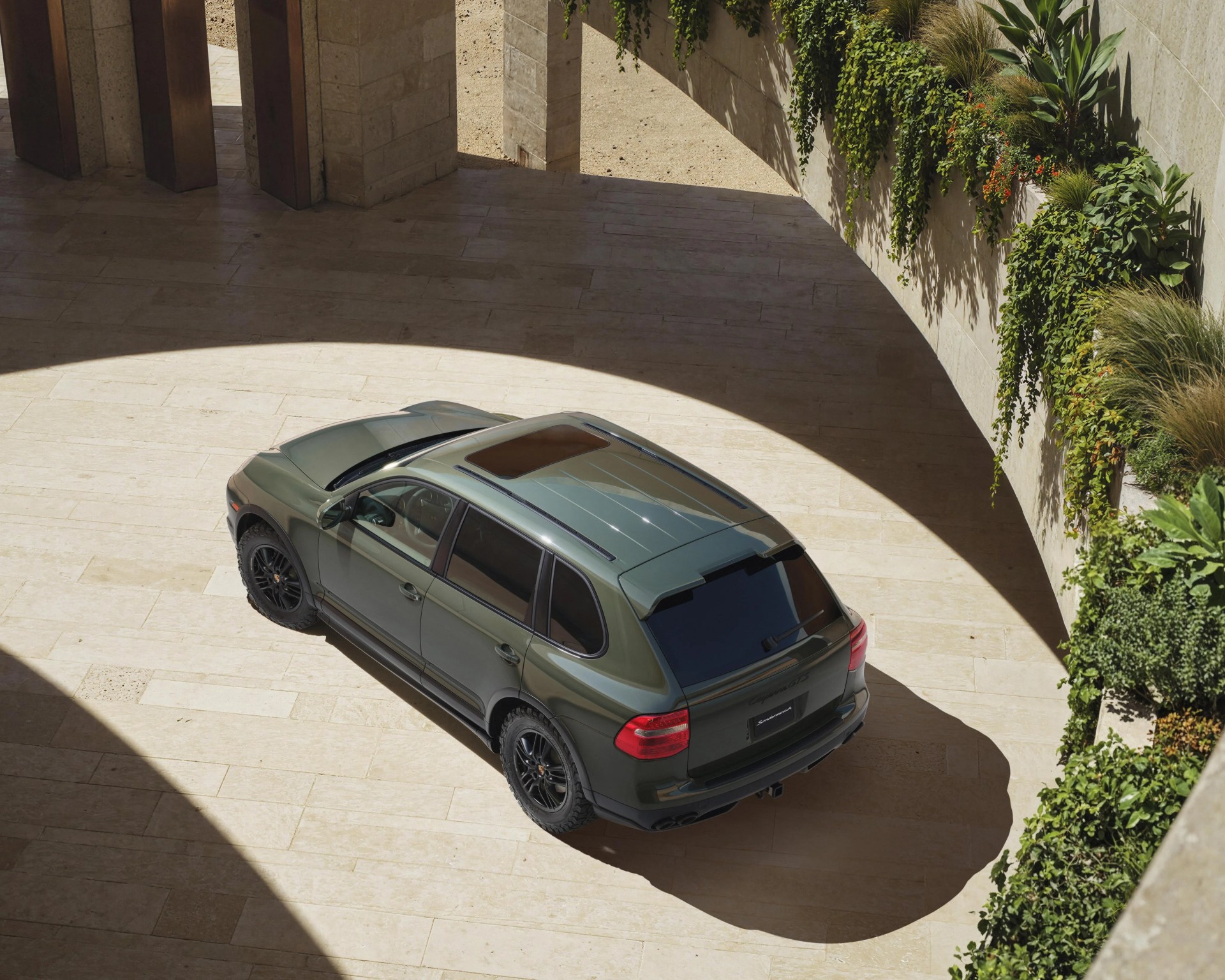 Green Porsche Cayenne SUV with black wheels parked on a light stone floor near a curved wall with hanging plants.