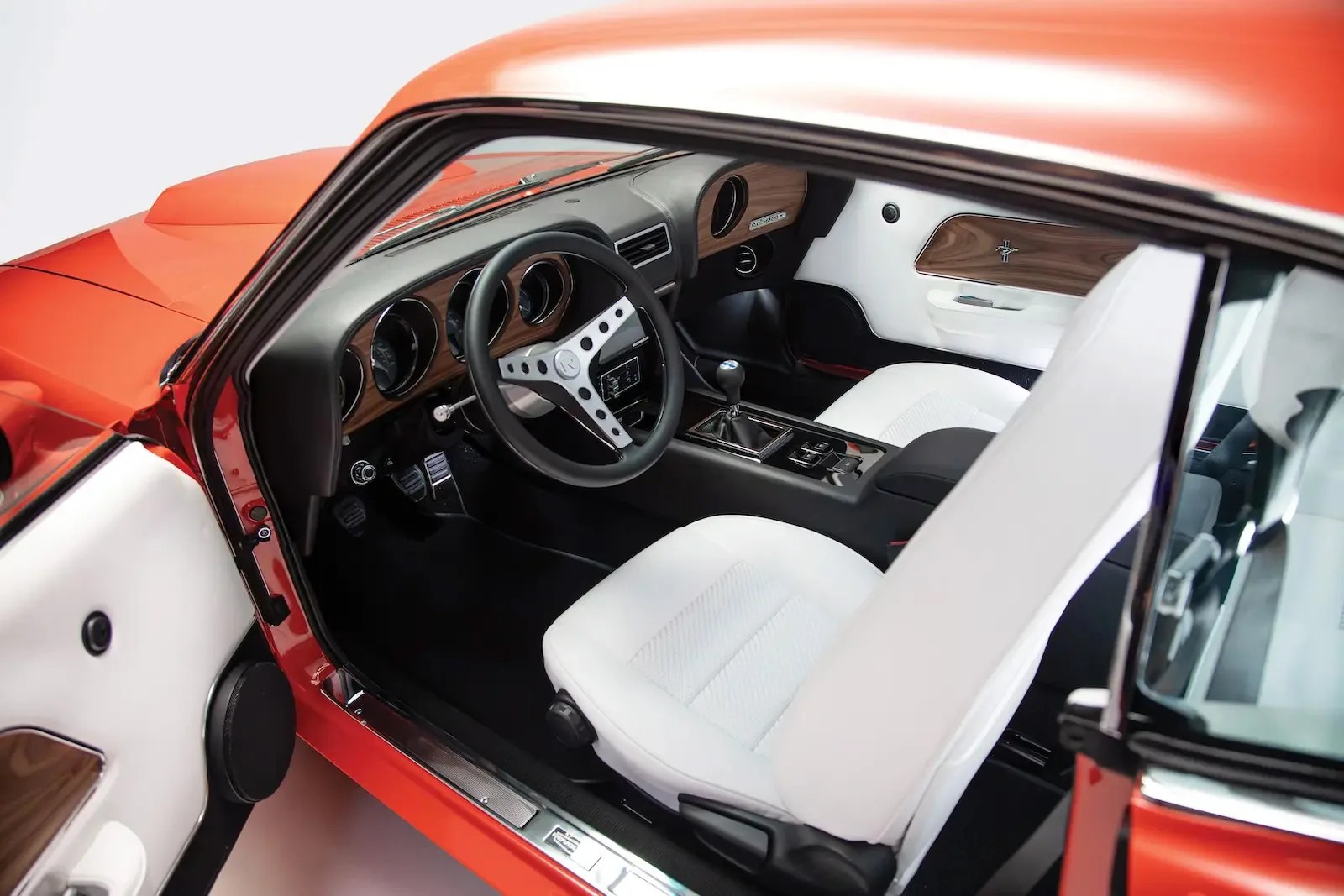 Interior of a classic car with white seats, wood panel dashboard, black steering wheel, and manual gear shift.