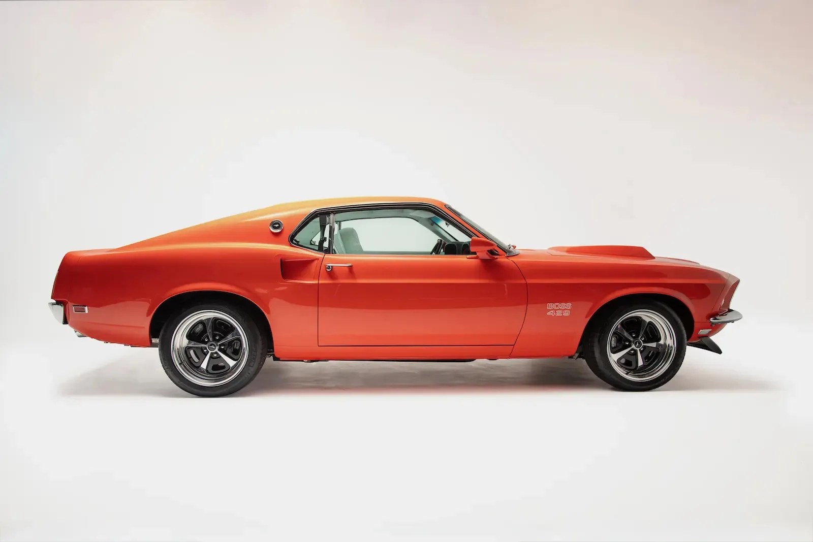 Orange 1969 Ford Mustang Boss 429 coupe with black five-spoke wheels on a white background.