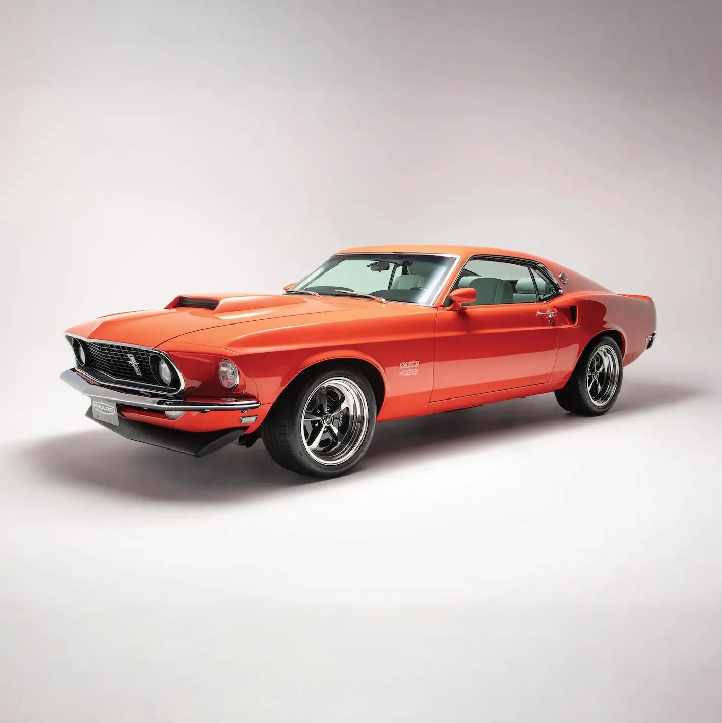 Orange 1969 Ford Mustang Boss 429 with black racing wheels on a white studio background.