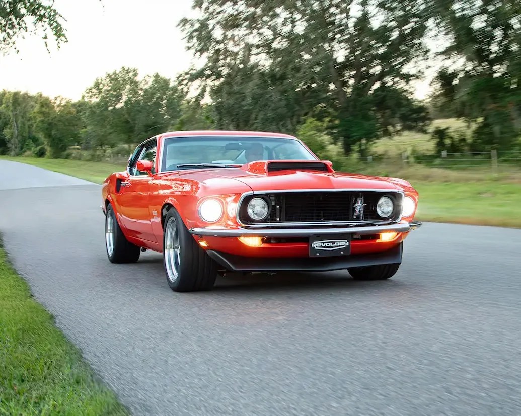 Red classic Ford Mustang with headlights on driving on a rural road with trees in the background.