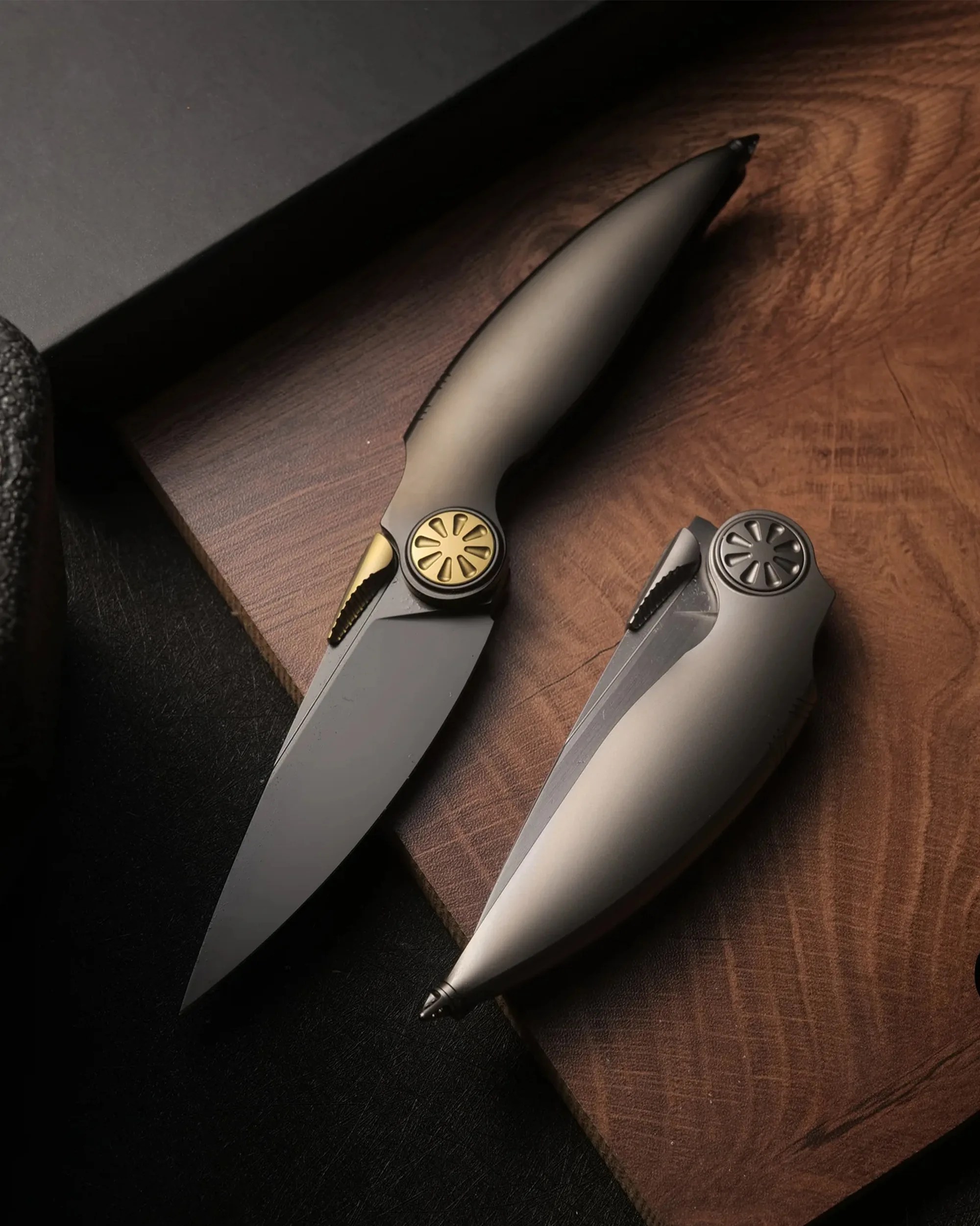 Two sleek folding knives with matte gray handles and gold and black accents on a wooden surface.
