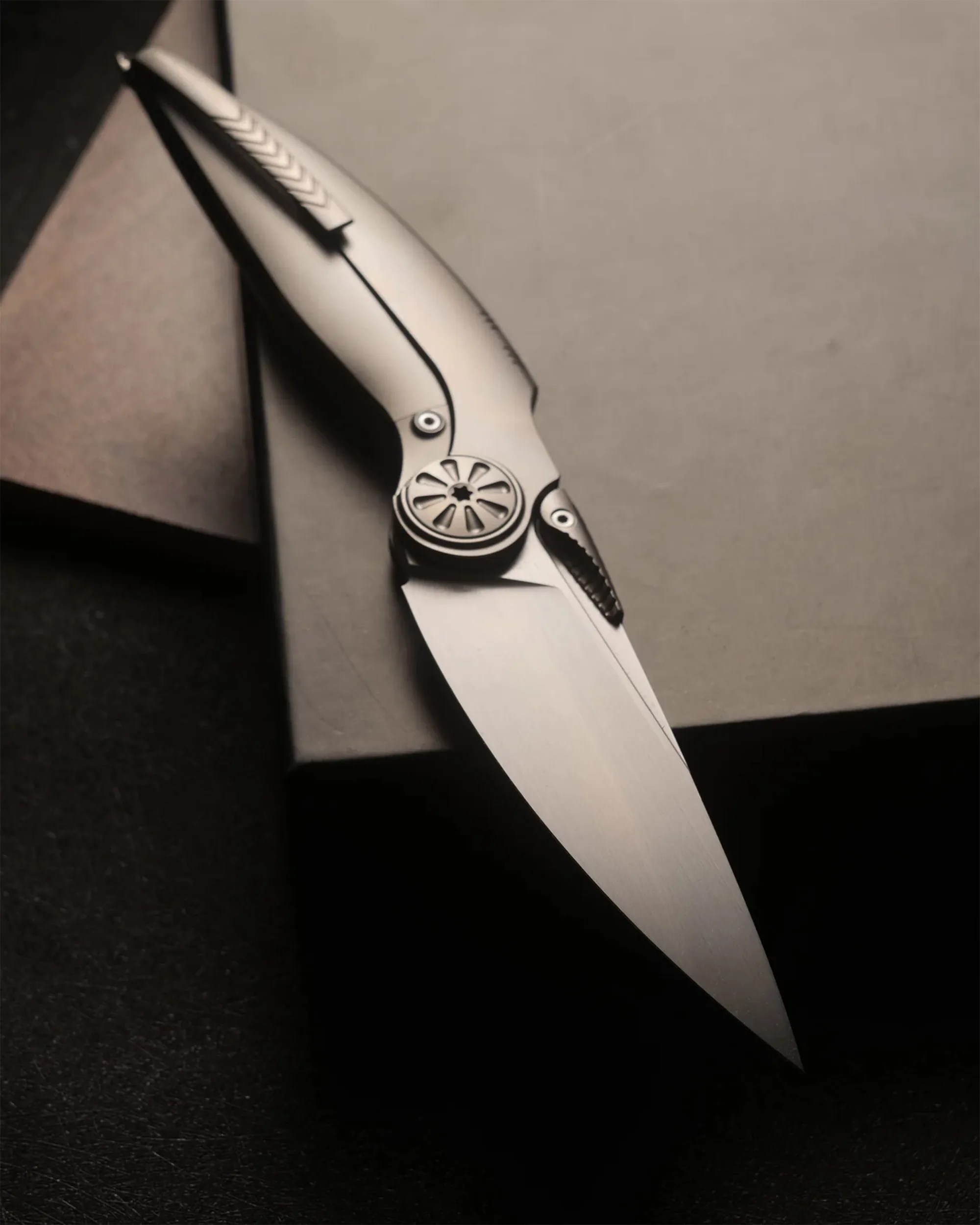 Sleek metallic folding knife with a smooth blade and intricate circular pivot design resting on a dark box.
