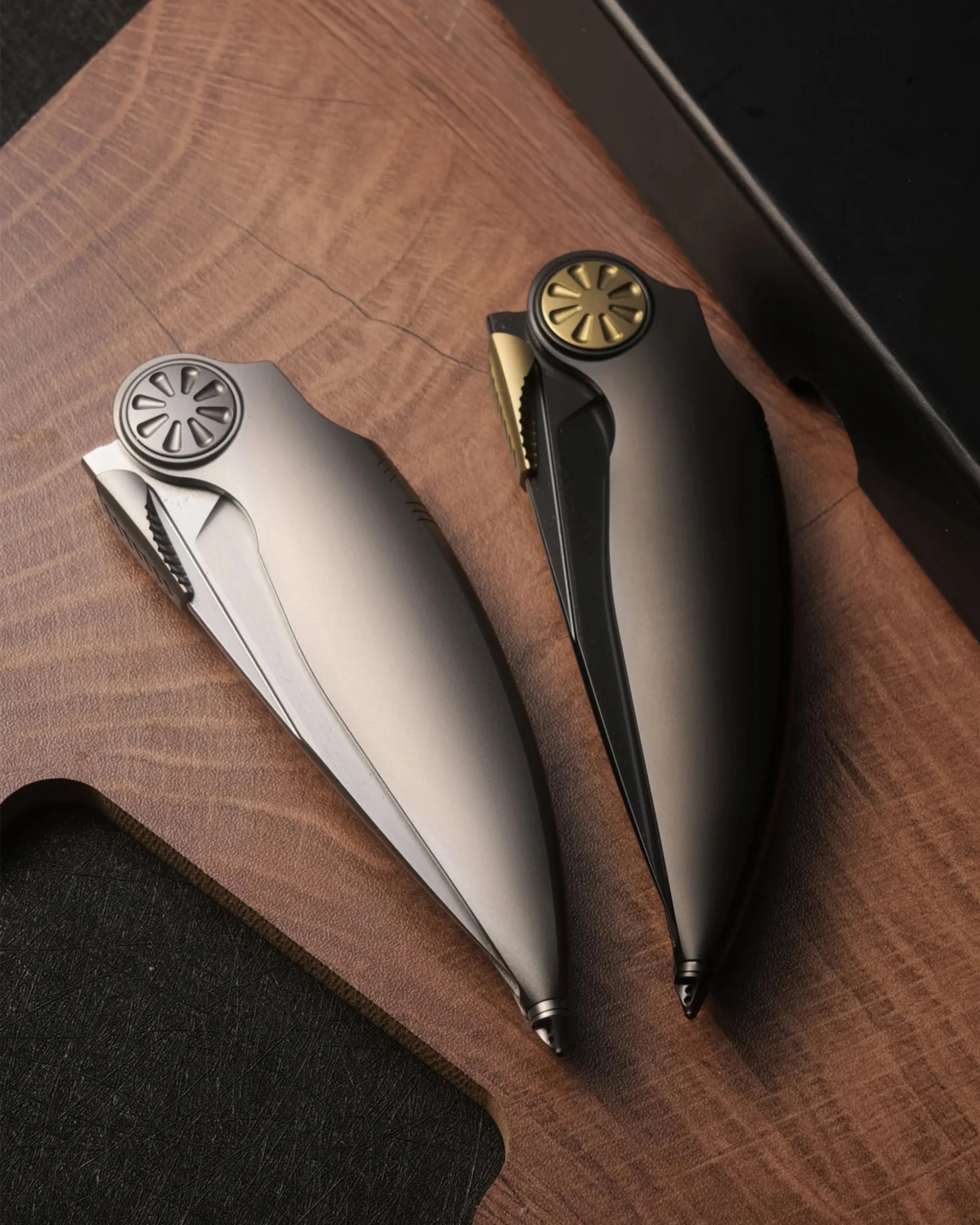 Two sleek folding knives with metallic finishes, one silver and one black with gold accents, on a wooden surface.