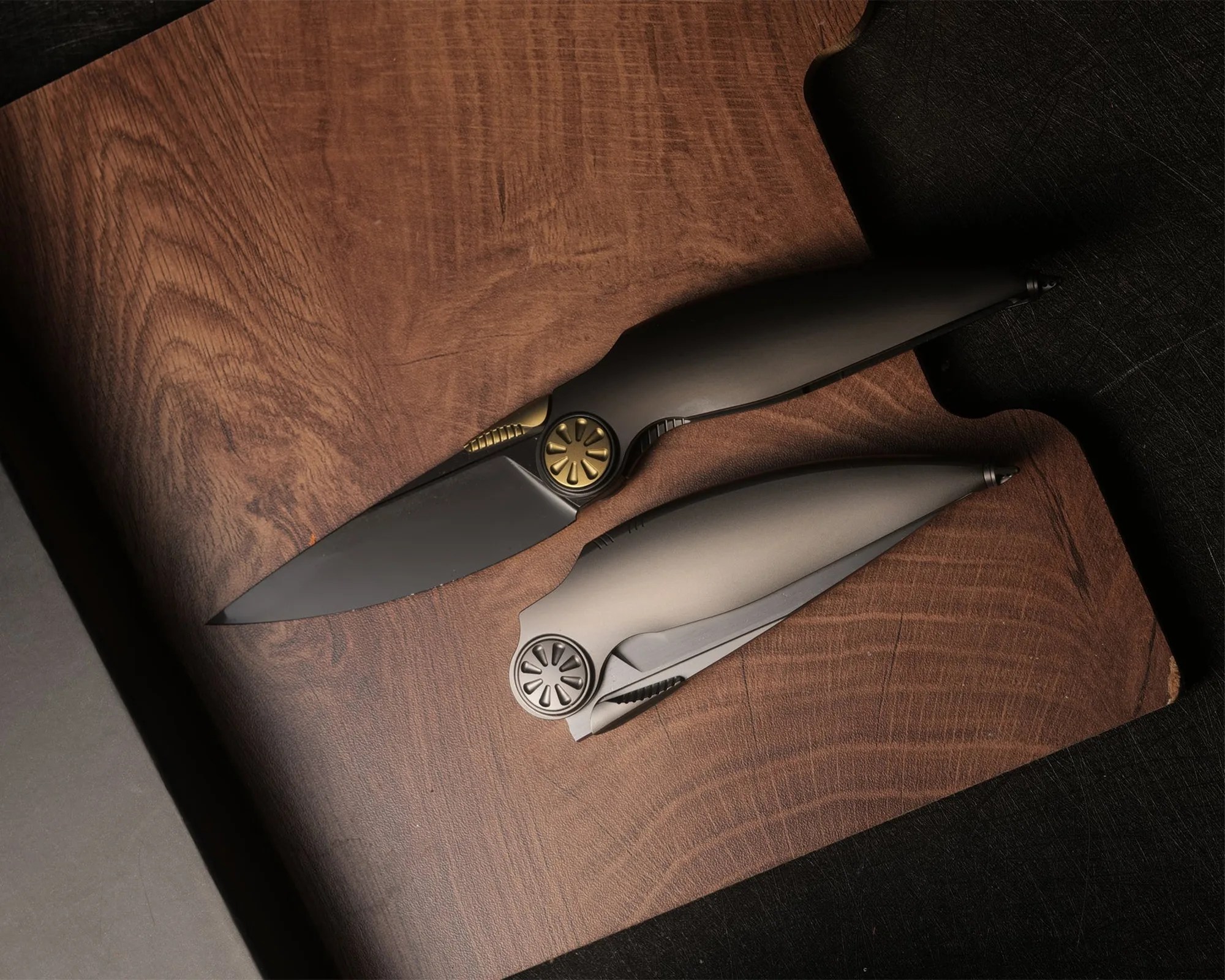 Two sleek folding knives, one open with a black blade and gold pivot, the other closed with a silver handle, on a wooden board.
