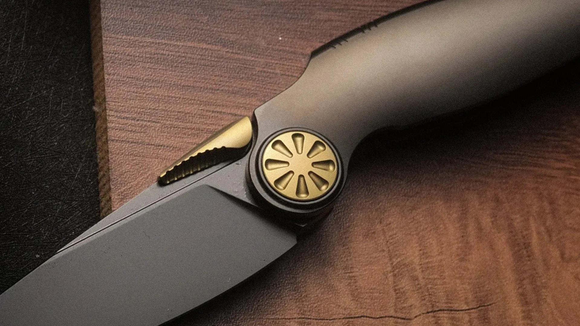 Close-up of a black folding knife with a matte blade and gold circular pivot with teardrop cutouts on a wooden surface.