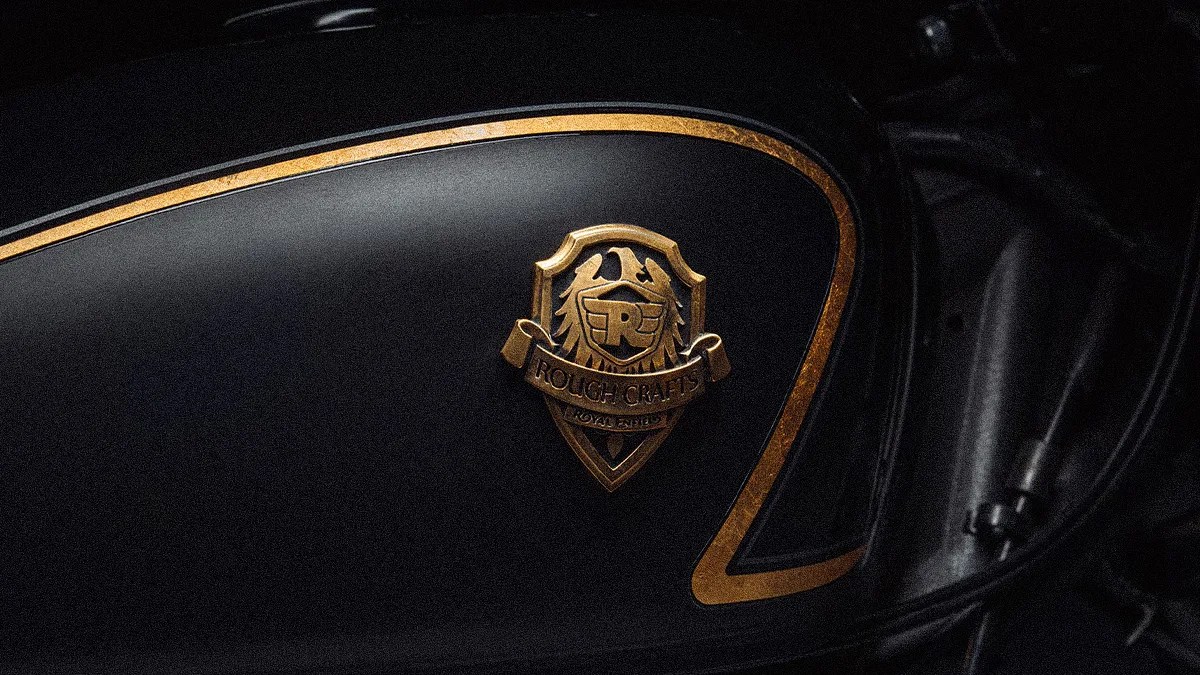 Black motorcycle fuel tank with gold trim and a gold "Rough Crafts Royal Enfield" emblem.