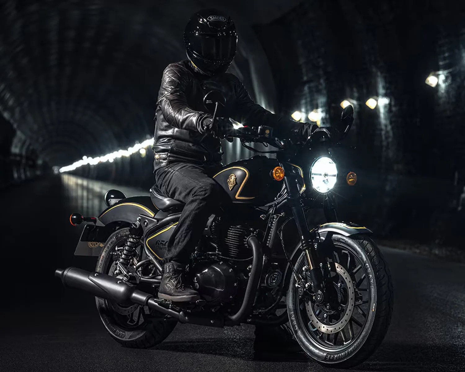 Black motorcycle with gold trim and bright headlight ridden by a person in black leather jacket and helmet inside a tunnel.