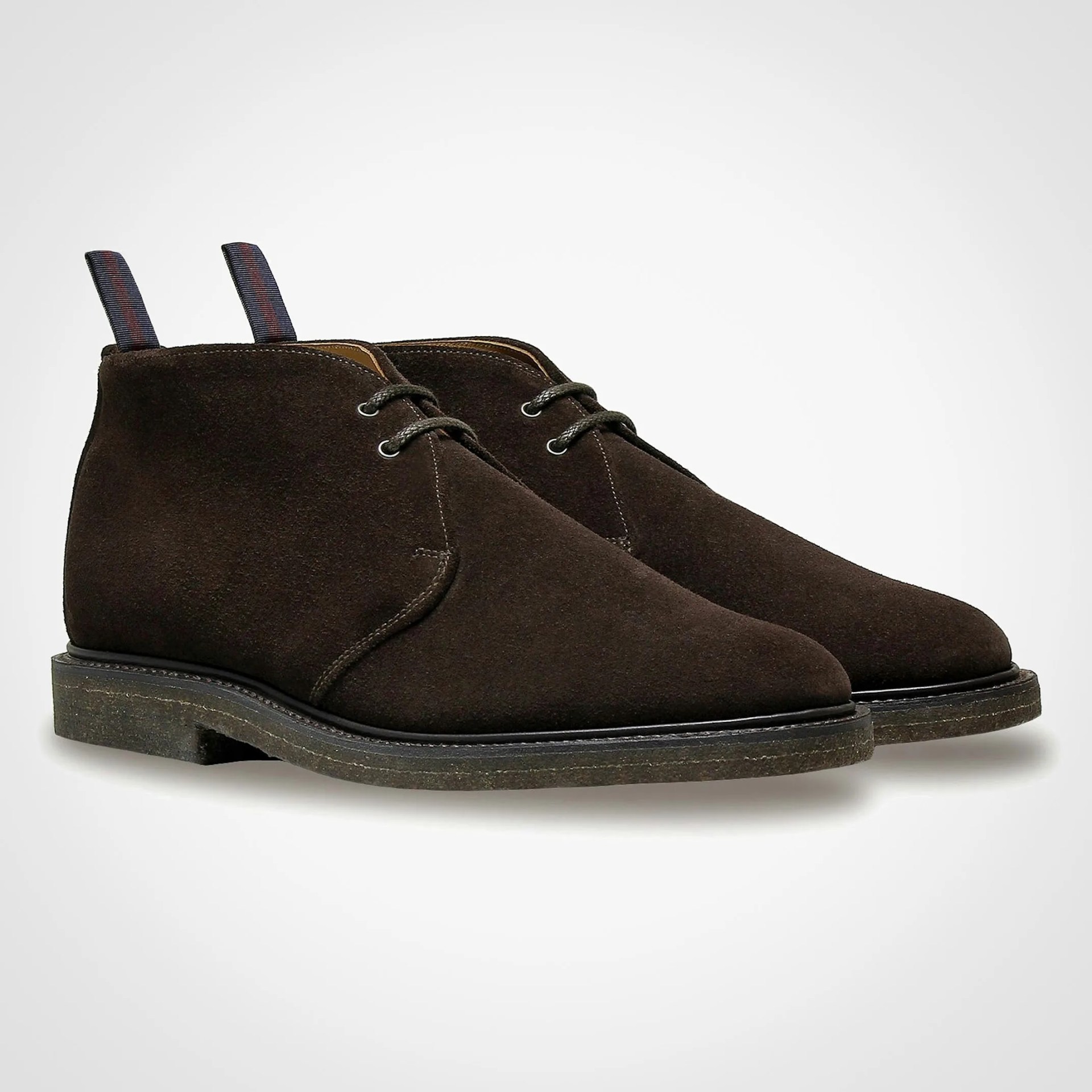 Pair of dark brown suede chukka boots with black soles and striped pull tabs at the back.