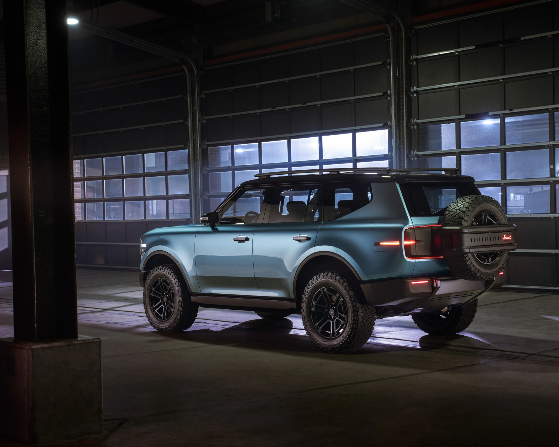 Matte blue-green SUV with black off-road tires and spare tire mounted on rear, parked inside a garage.