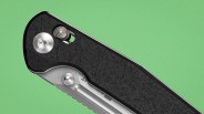 Close-up of a black textured folding knife handle with visible metal screws and part of the blade against a green background.