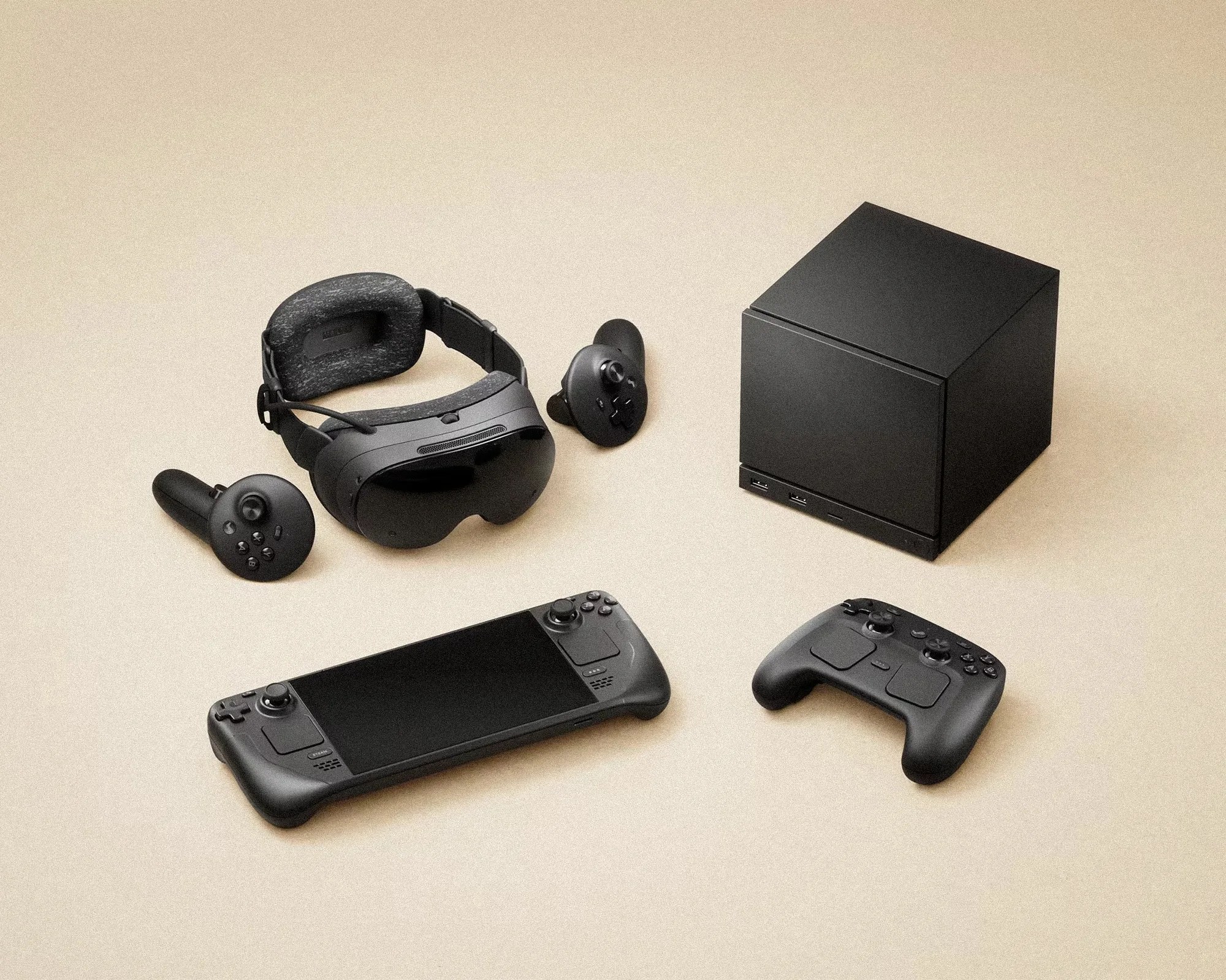 Black VR headset with two controllers, a black cube-shaped PC, a handheld gaming console, and a game controller on a beige surface.