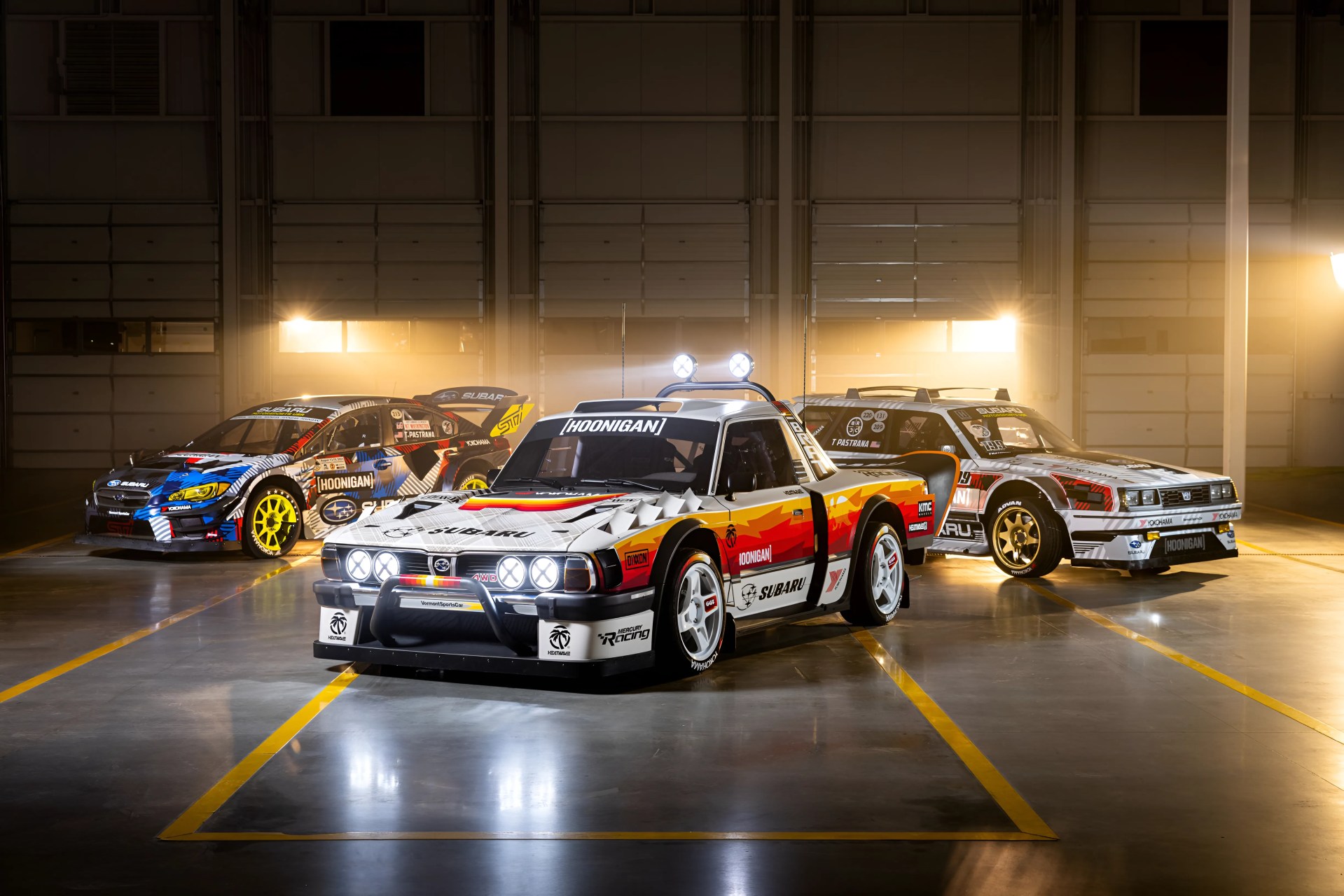 Three Subaru rally cars with colorful racing decals and bright headlights in a dimly lit garage.