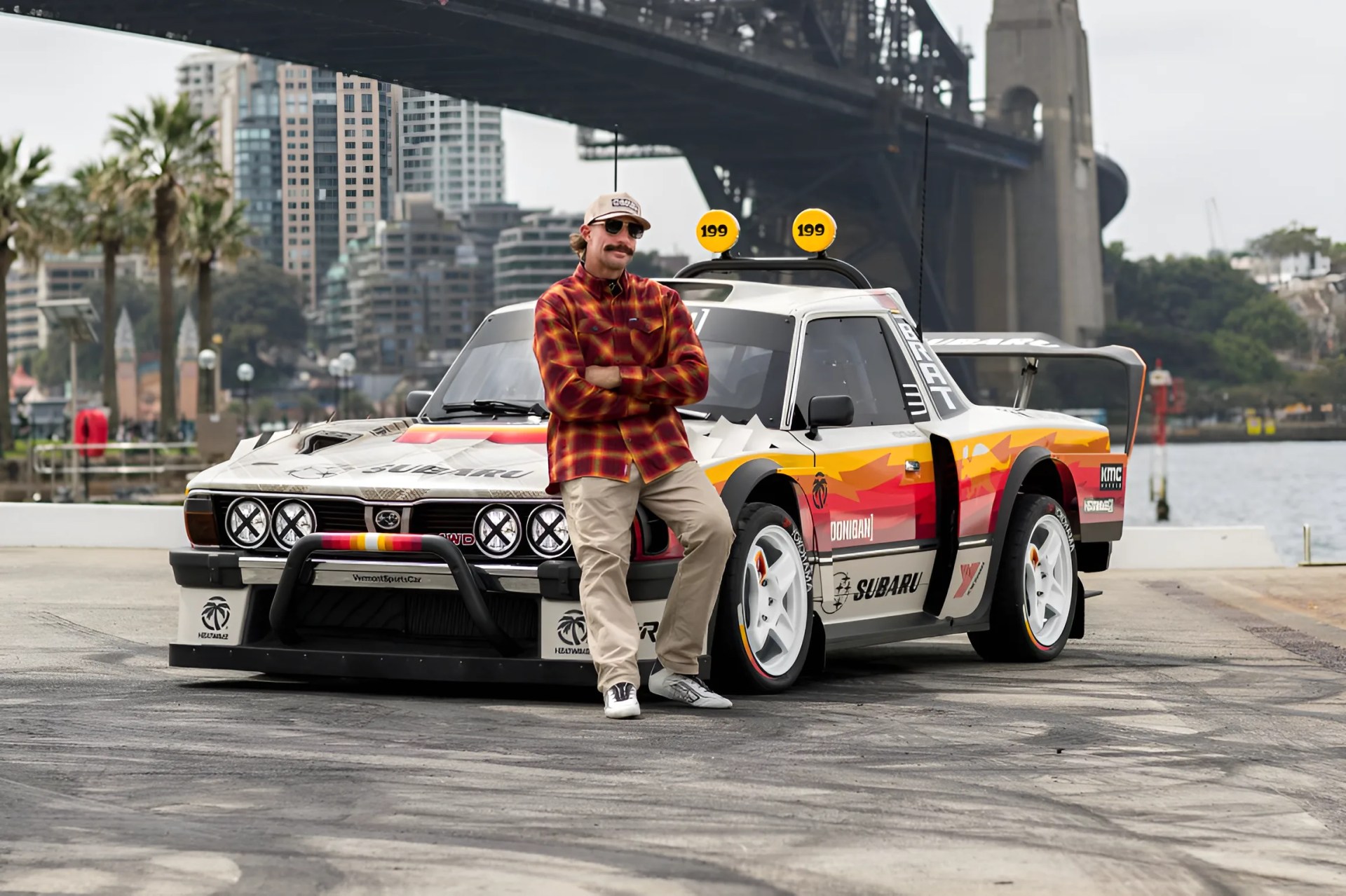 Man in a red plaid shirt and beige pants leaning against a white Subaru rally car with colorful decals under a bridge.