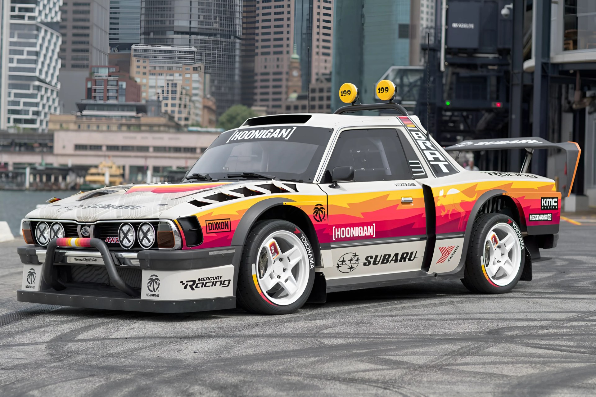 White and orange Subaru BRAT race car with Hoonigan branding and roof-mounted yellow lights parked on asphalt.