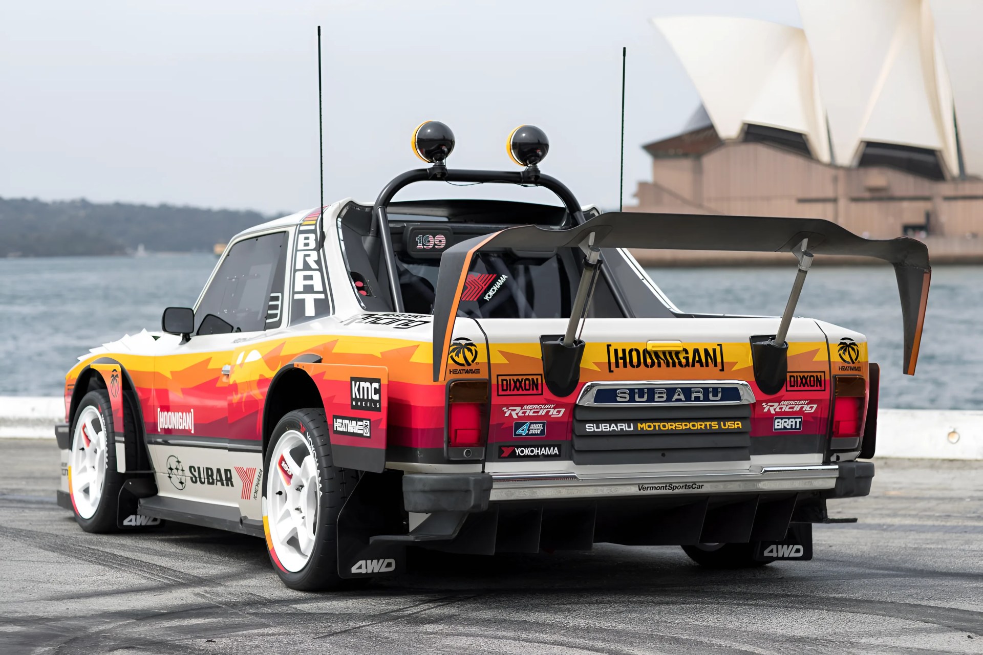 White Subaru BRAT race car with red and orange gradient decals, large rear wing, and white wheels near water.