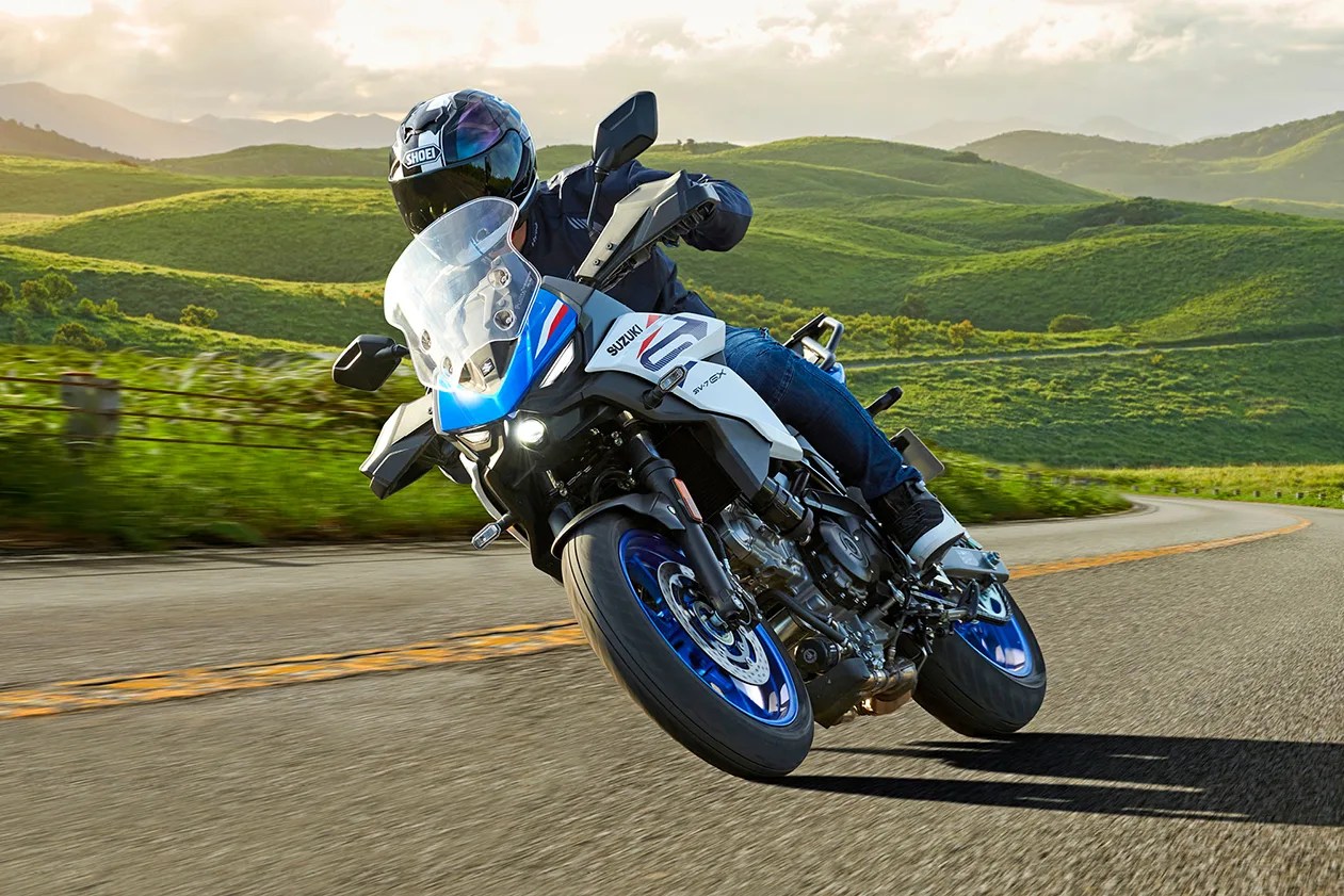 Rider wearing a black helmet and jacket leaning into a turn on a white and blue Suzuki SV 650X motorcycle on a winding road with green hills in the background.