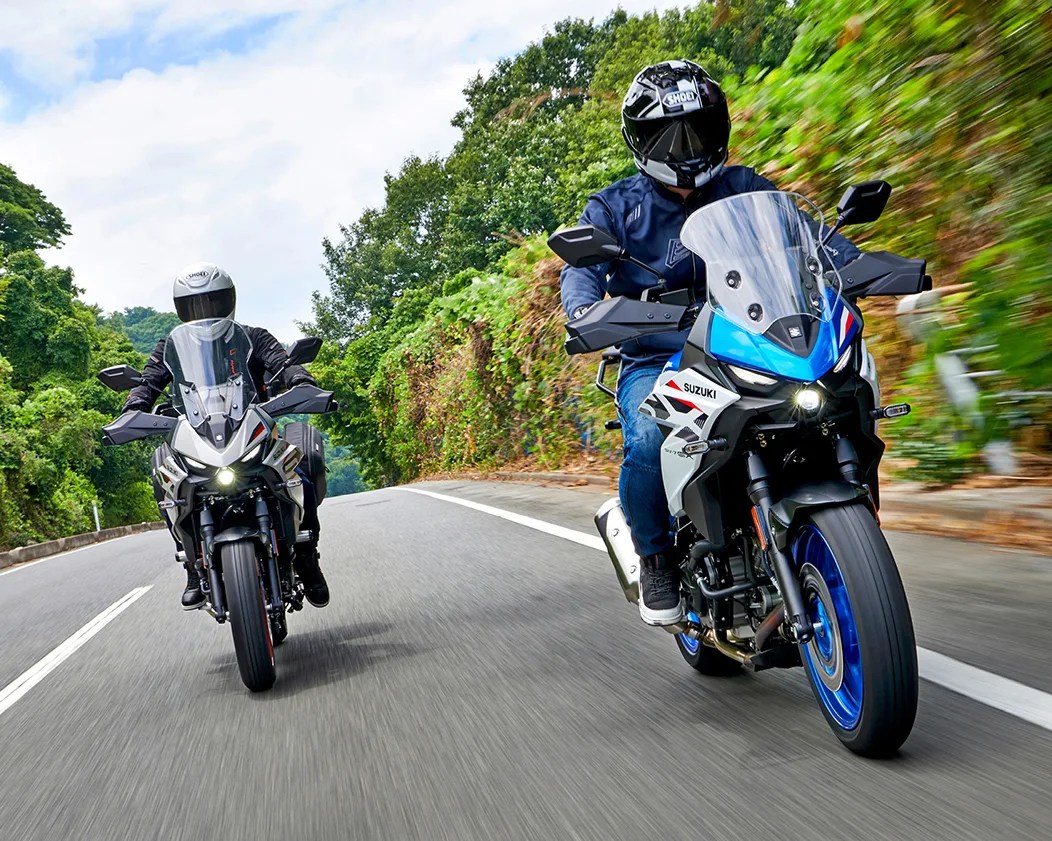 Two riders wearing helmets on Suzuki adventure motorcycles riding on a paved road with greenery on the sides.