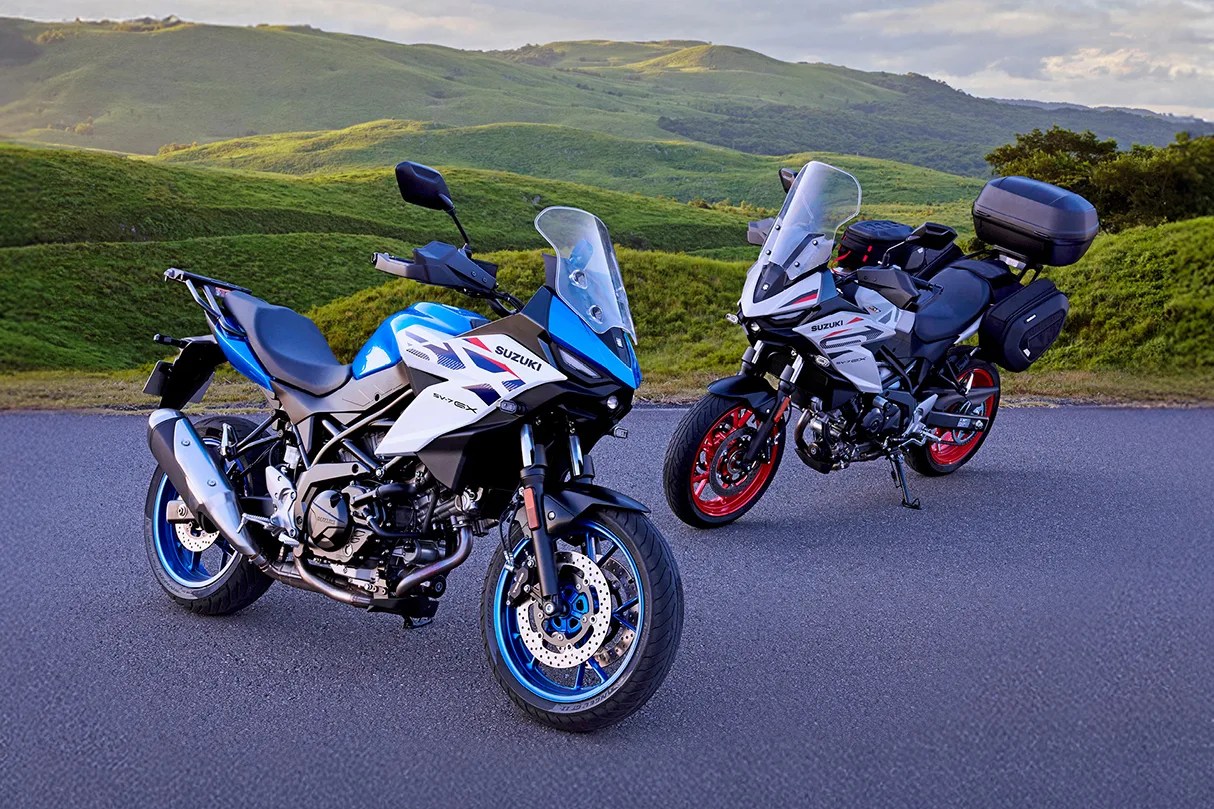Two Suzuki SV650X motorcycles, one blue and white with blue rims and one white with red rims and saddlebags, parked on a road with green hills in the background.
