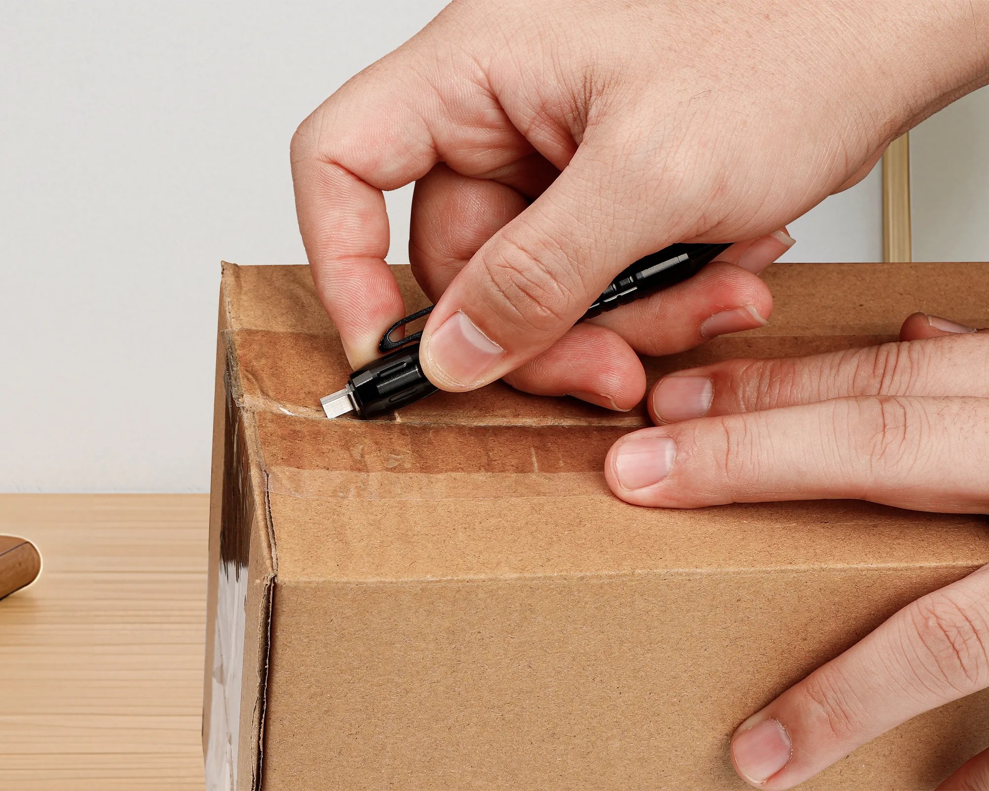 Hands using a small black utility knife to cut open a sealed cardboard box on a wooden surface.