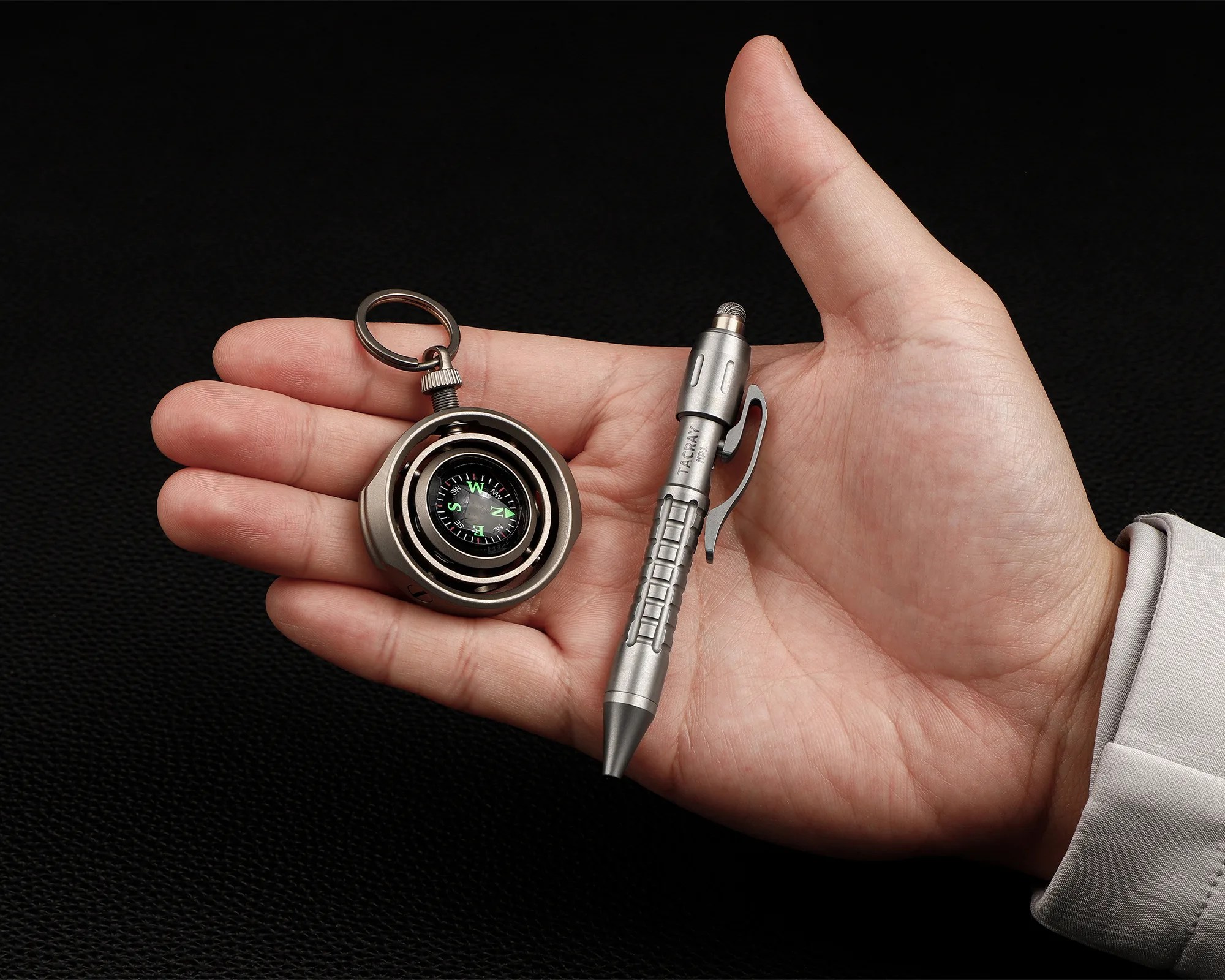 Hand holding a silver metal compass keychain and a silver tactical pen with a clip.