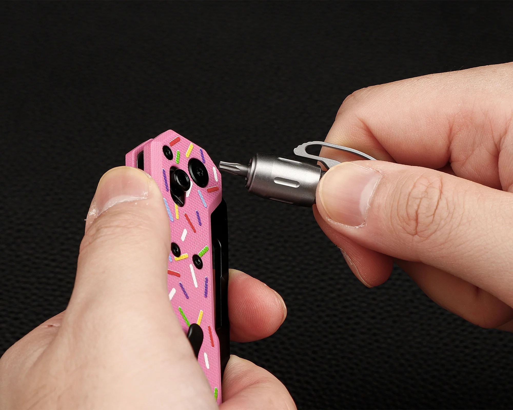 Hands holding a pink multi-tool with colorful sprinkles pattern and a small silver screwdriver bit.