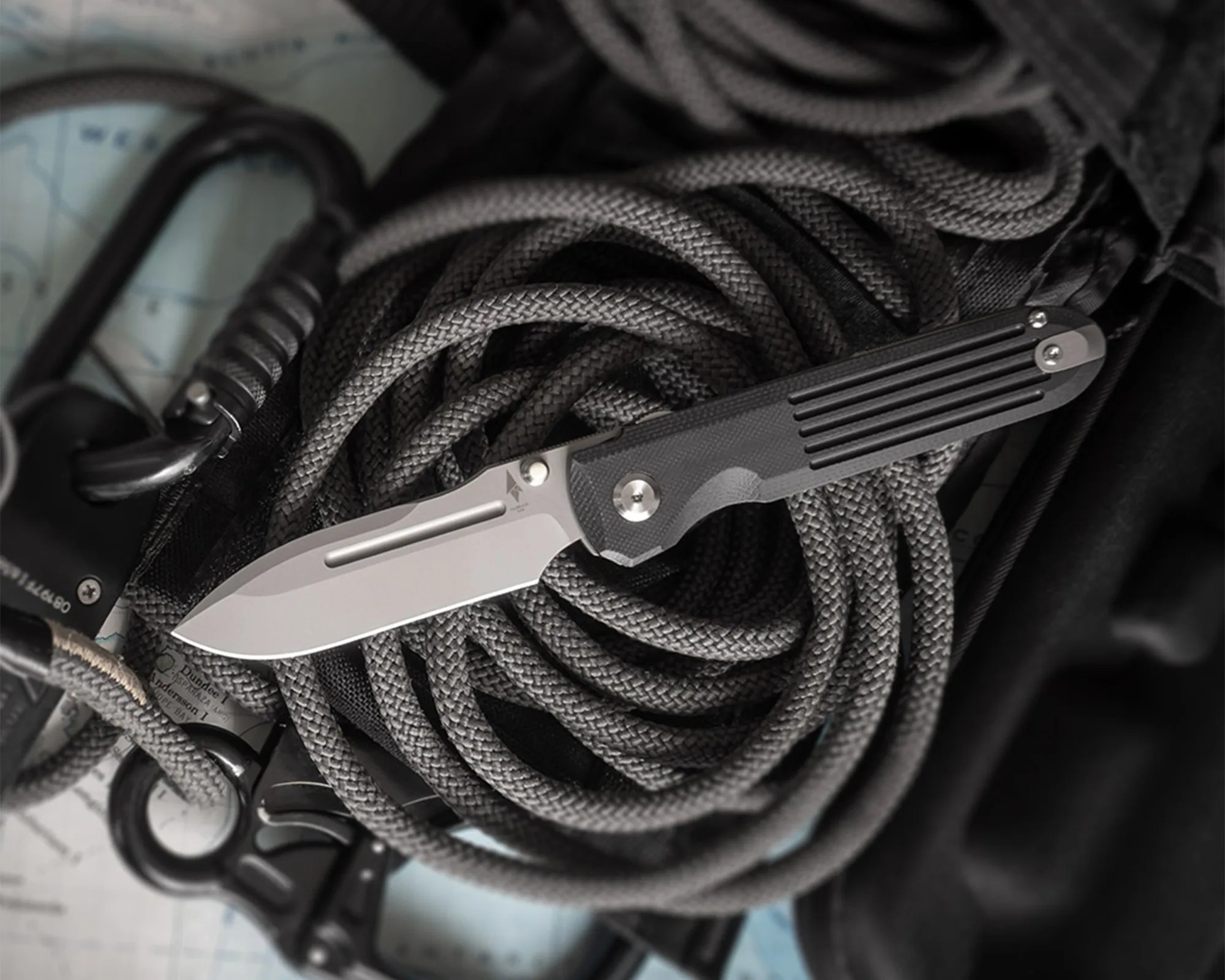 Folding knife with a silver blade and black textured handle resting on coiled gray rope and black carabiners.