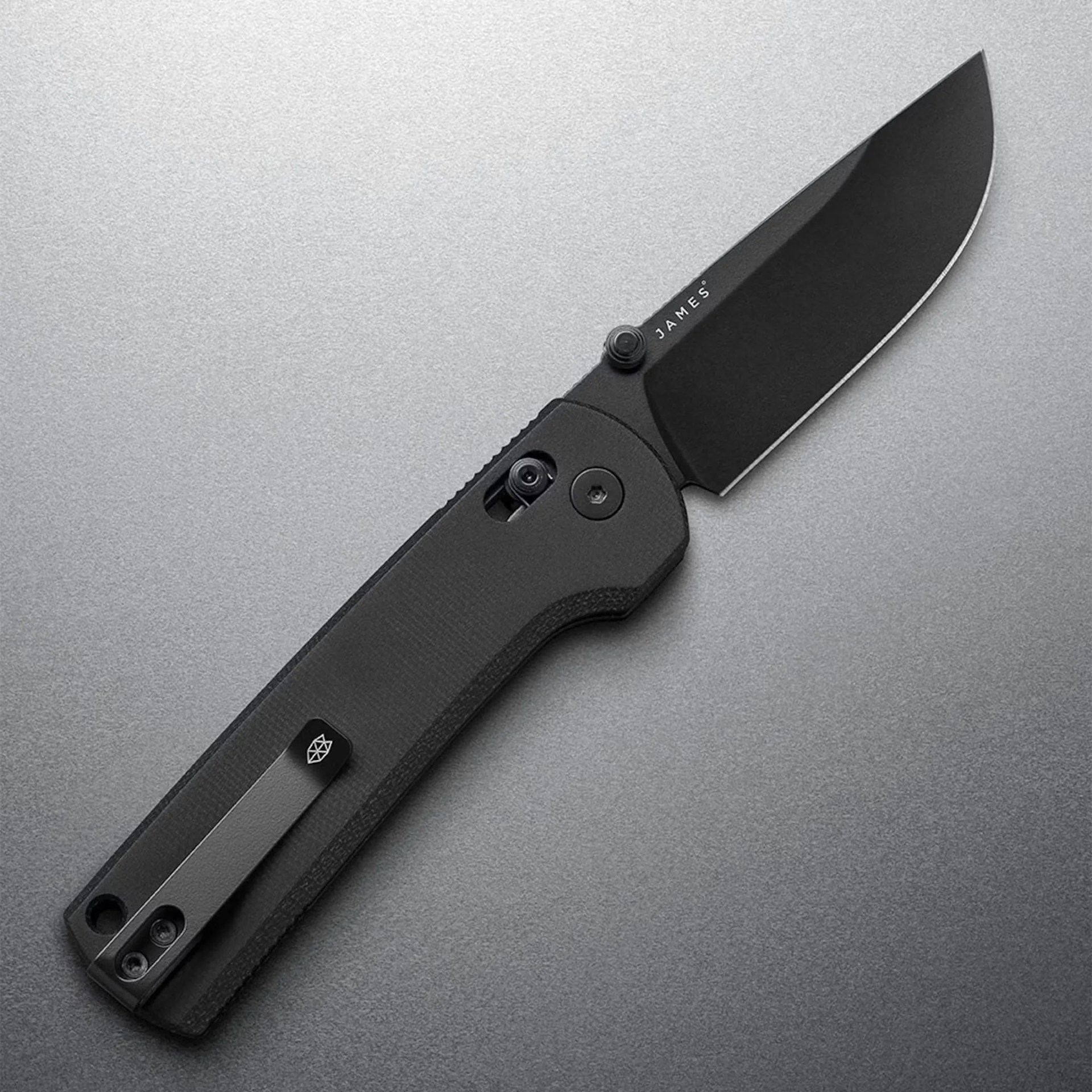 Black folding knife with textured handle and clip, blade marked "JAMES," on a gray surface.