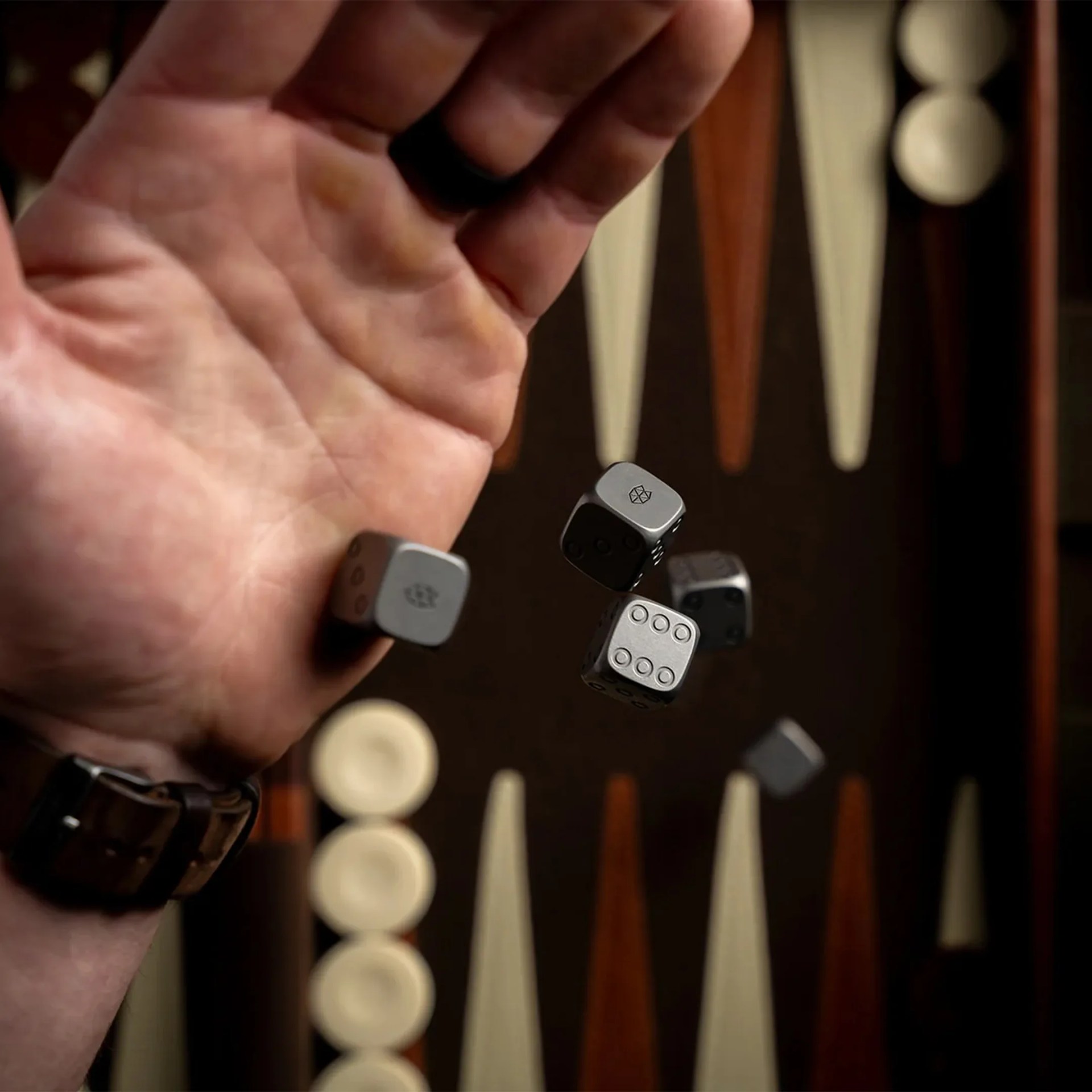 Hand wearing a dark watch rolling five metallic dice over a backgammon board.