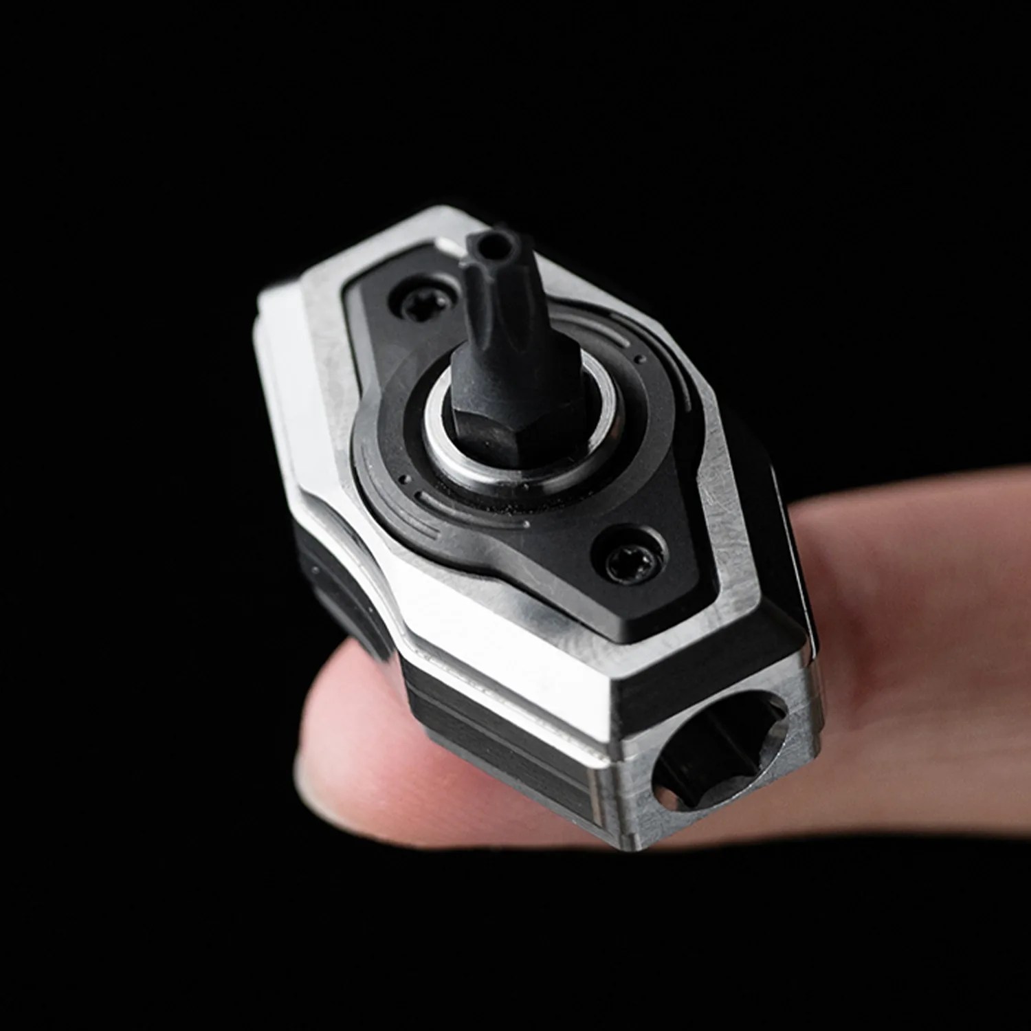 Compact metal and black plastic mechanical component held on a fingertip against a black background.