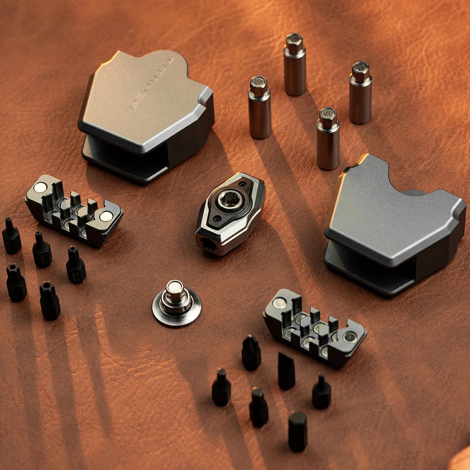 Two gray metal multi-bit screwdriver handles with multiple black screwdriver bits and four metal bit holders on a brown leather surface.