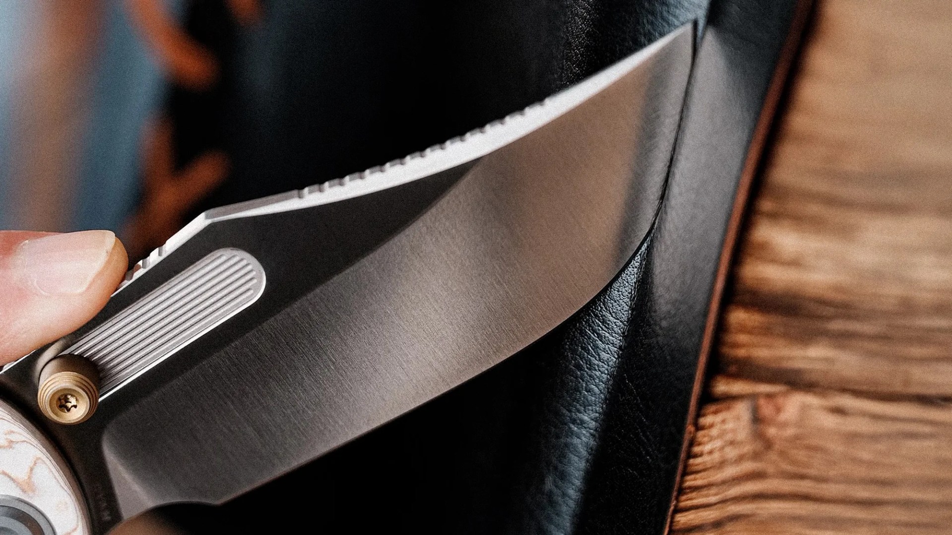 Close-up of a hand holding a folding knife with a partially serrated stainless steel blade against black leather and wooden surface.