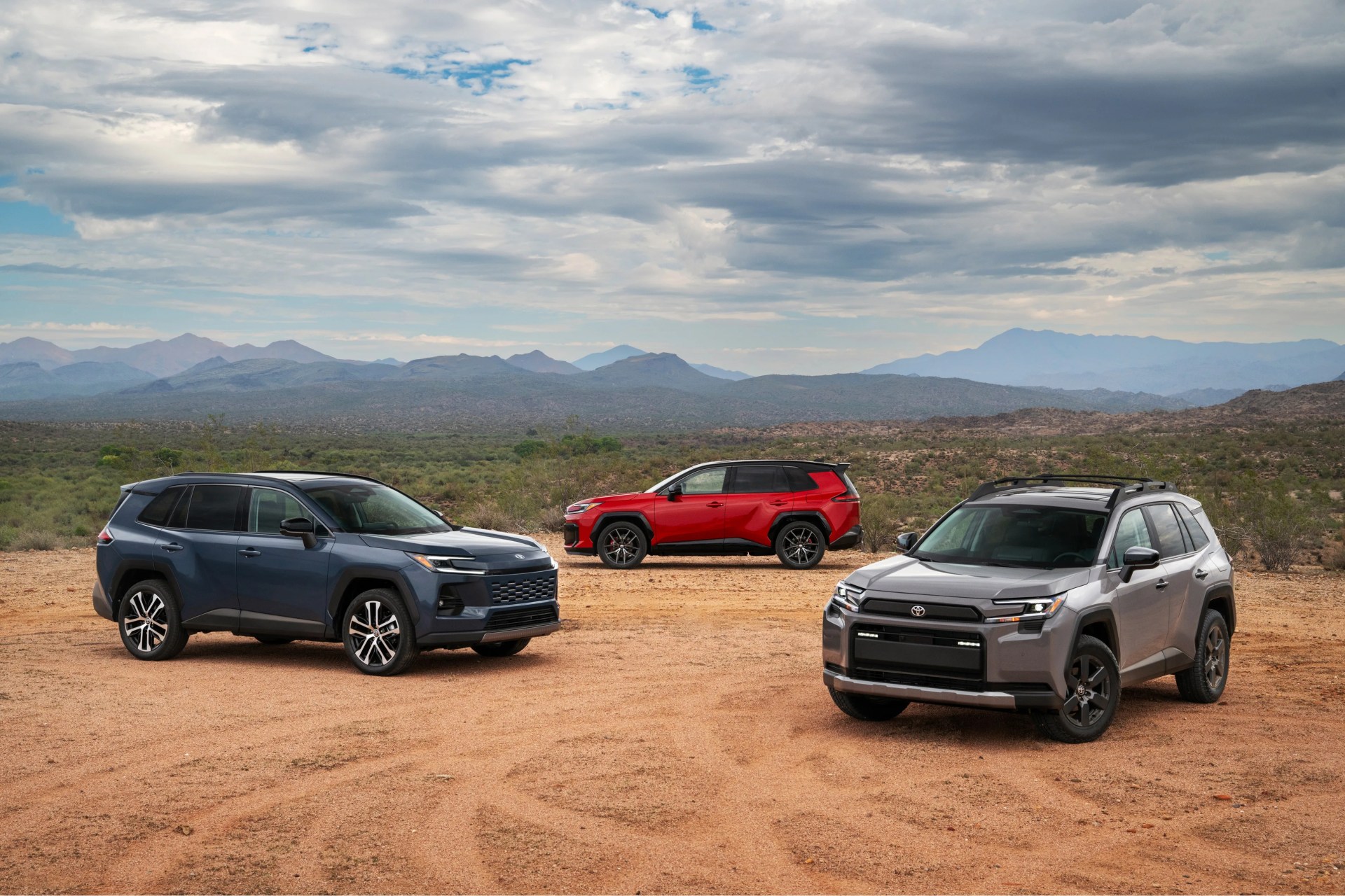Three mid-size SUVs in gray, blue, and red parked on a dirt area with mountains and cloudy sky in the background.