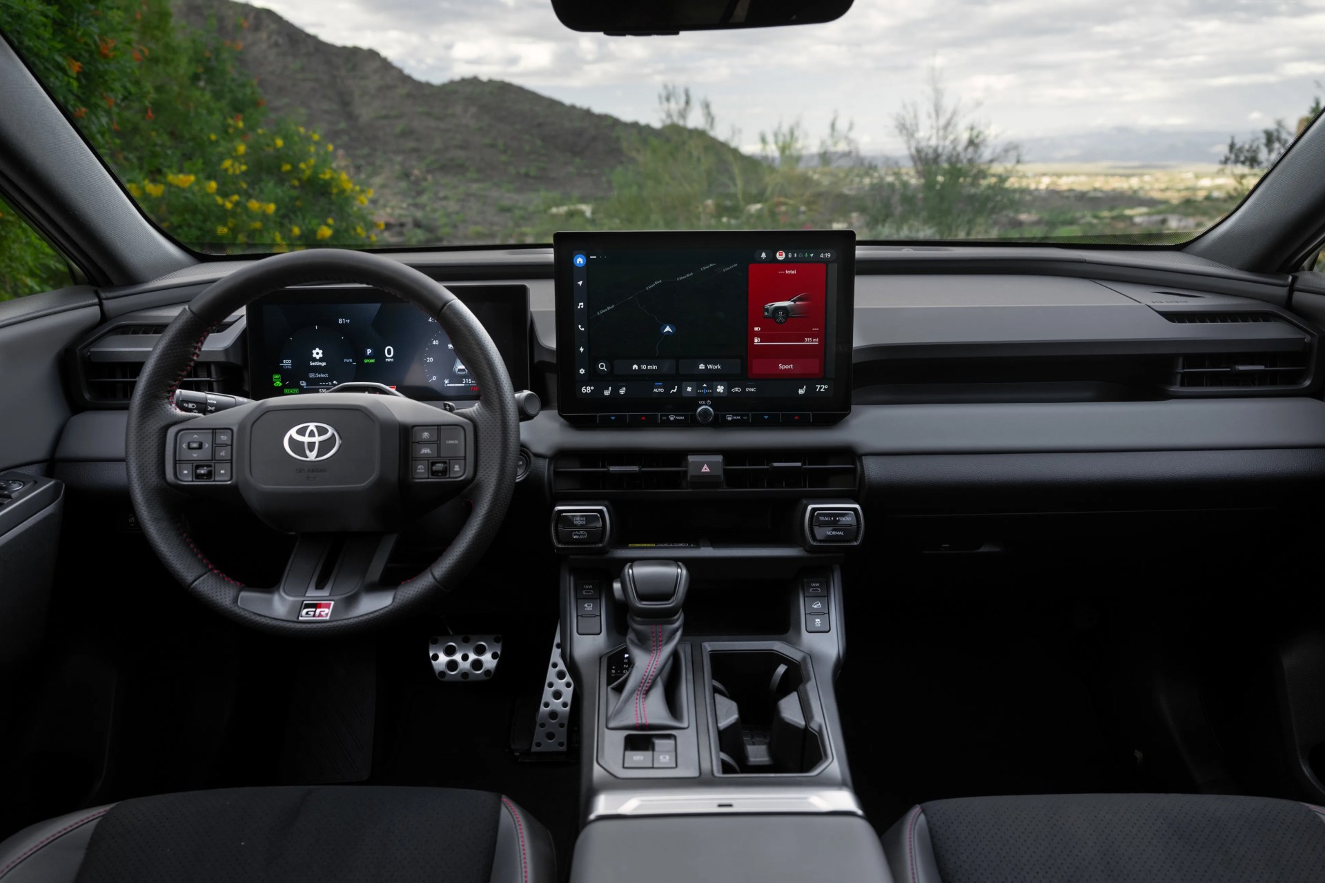 Black Toyota GR steering wheel and dashboard with digital displays and center touchscreen in a car interior.